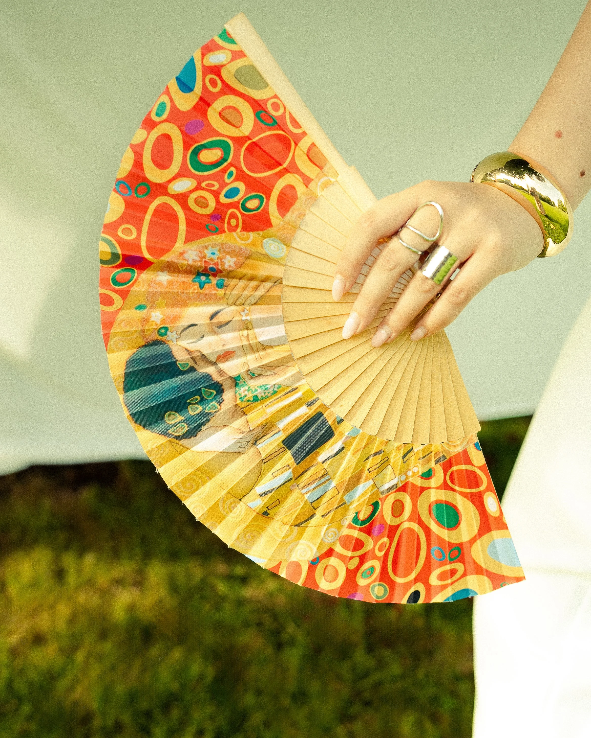 Person holding a colorful, decorated folding fan with rings and a gold bracelet on their wrist.