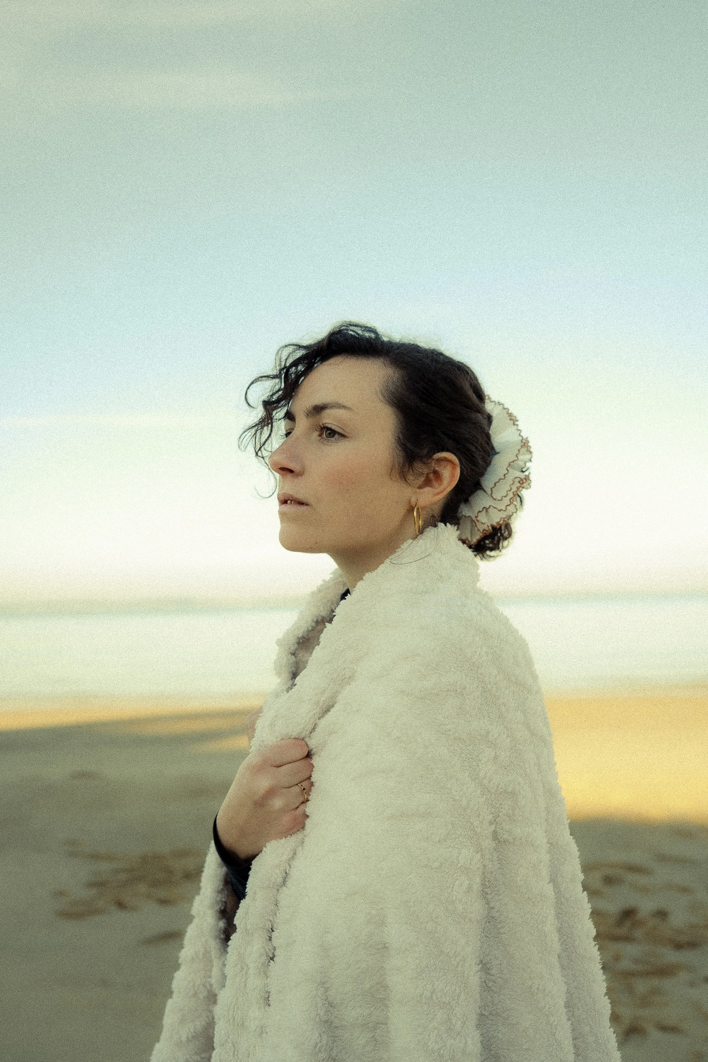 A woman with dark, curly hair wearing a cream-colored, textured coat at the beach, with a calm expression and the ocean in the background during sunset.