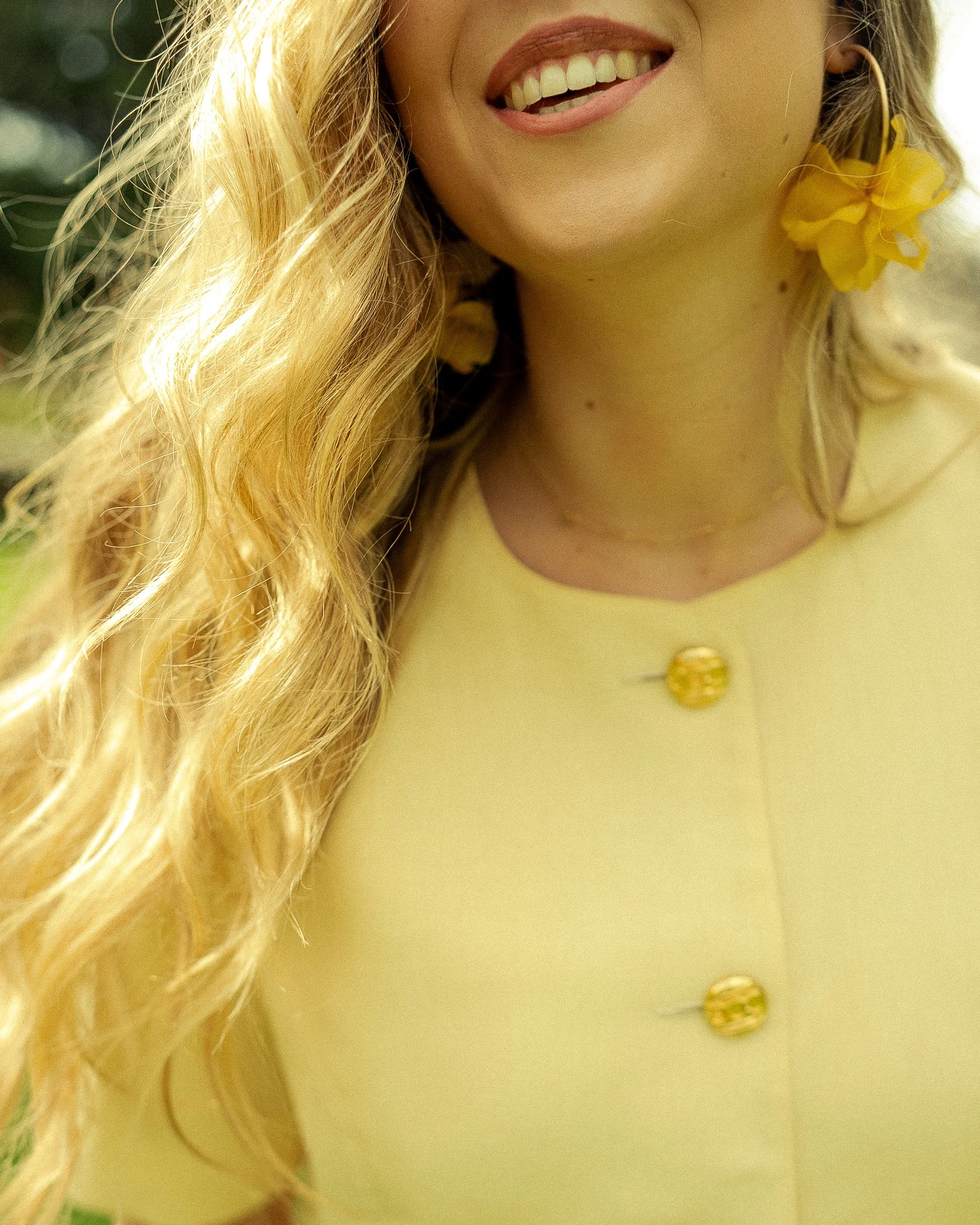 Close-up of a woman's smiling face, showing her teeth. She has long, wavy blonde hair, and is wearing yellow lipstick, yellow earrings with large flowers, and a yellow top with gold buttons.