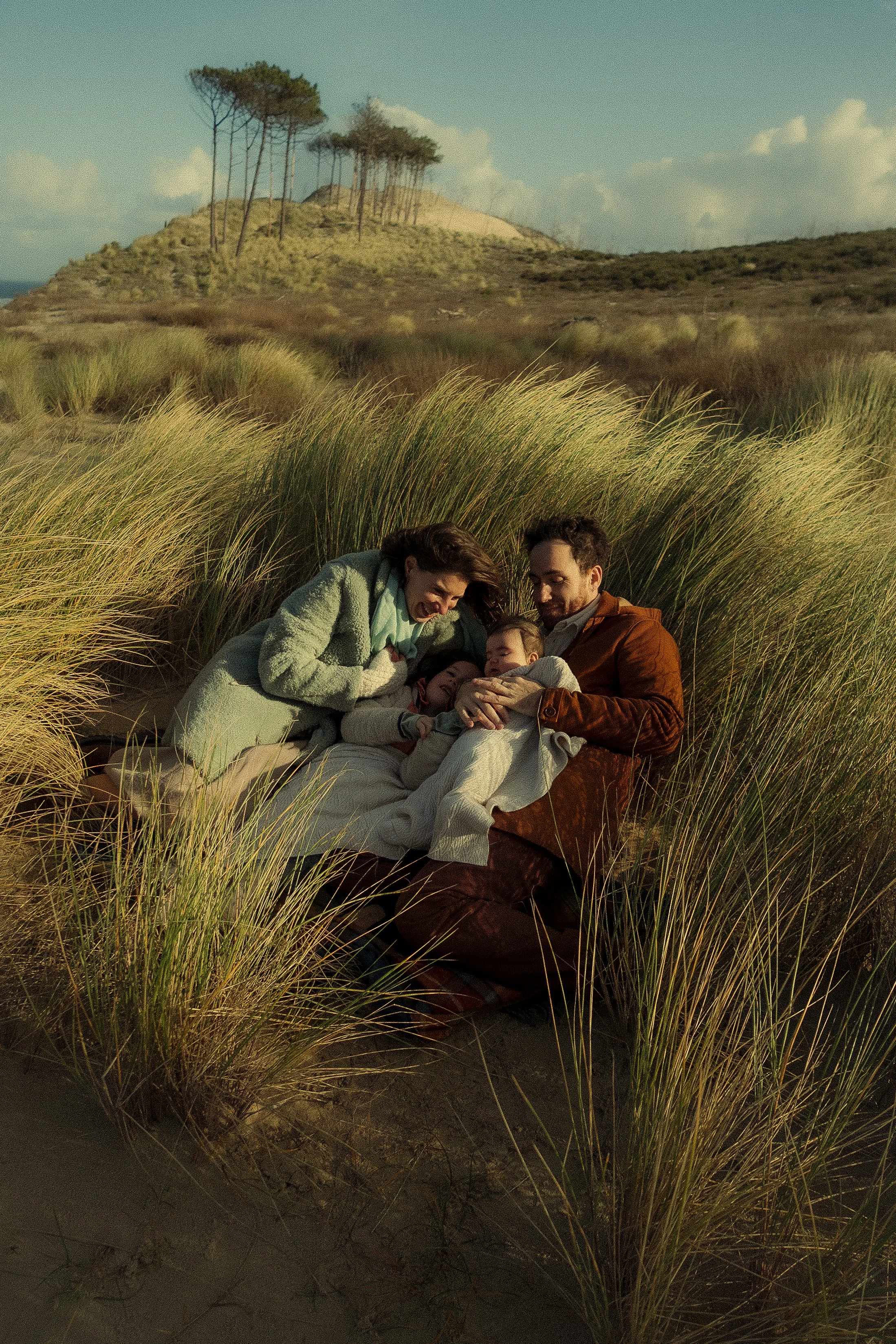 Une famille de quatre personnes, deux adultes et deux enfants, assise dans un champ d'herbes hautes, partageant un moment de tendresse en plein air, avec une colline et quelques arbres au loin.