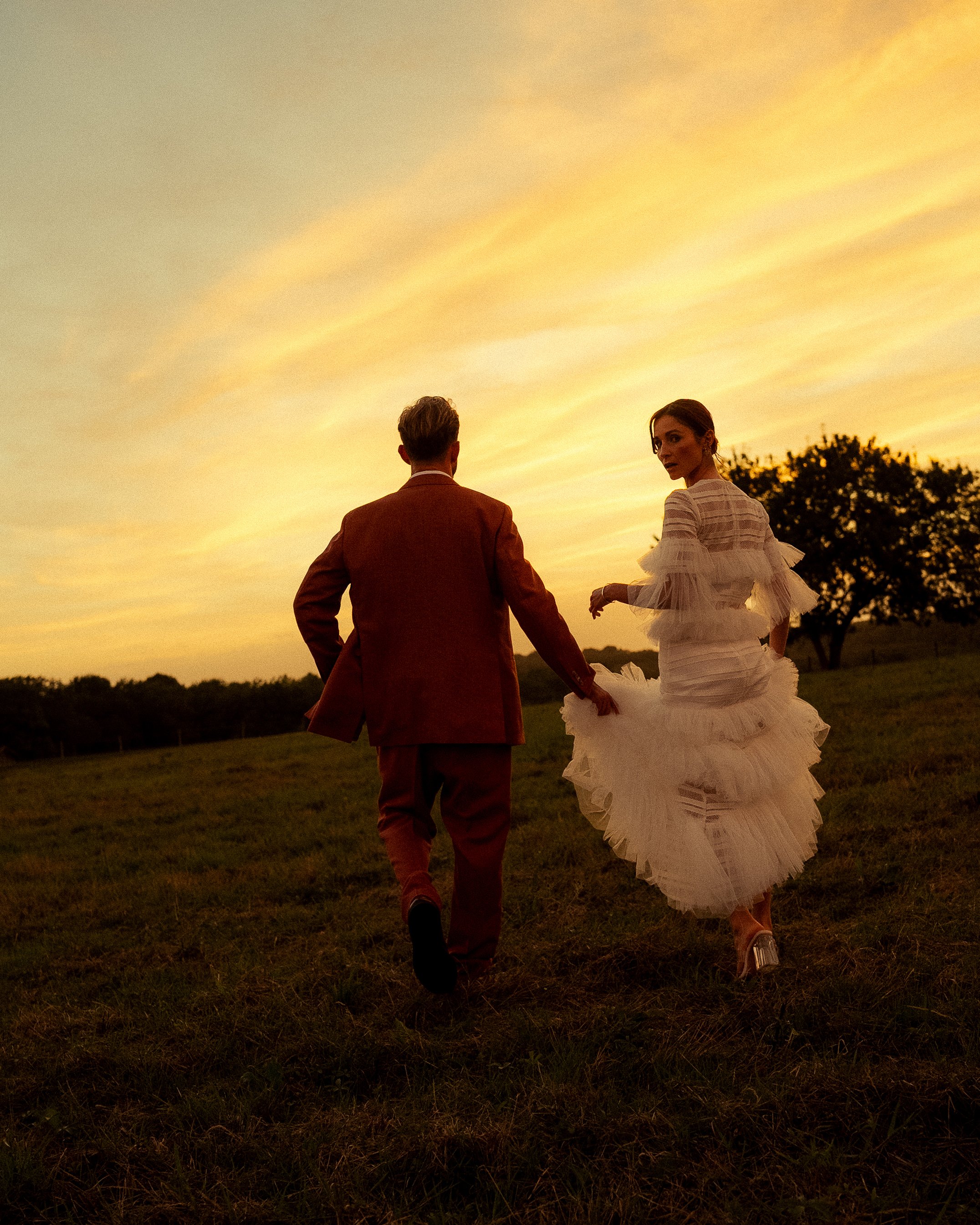 un couple marié marche dans un champ face au couché de soleil orange, l'homme tient d'une main la robe de sa femme