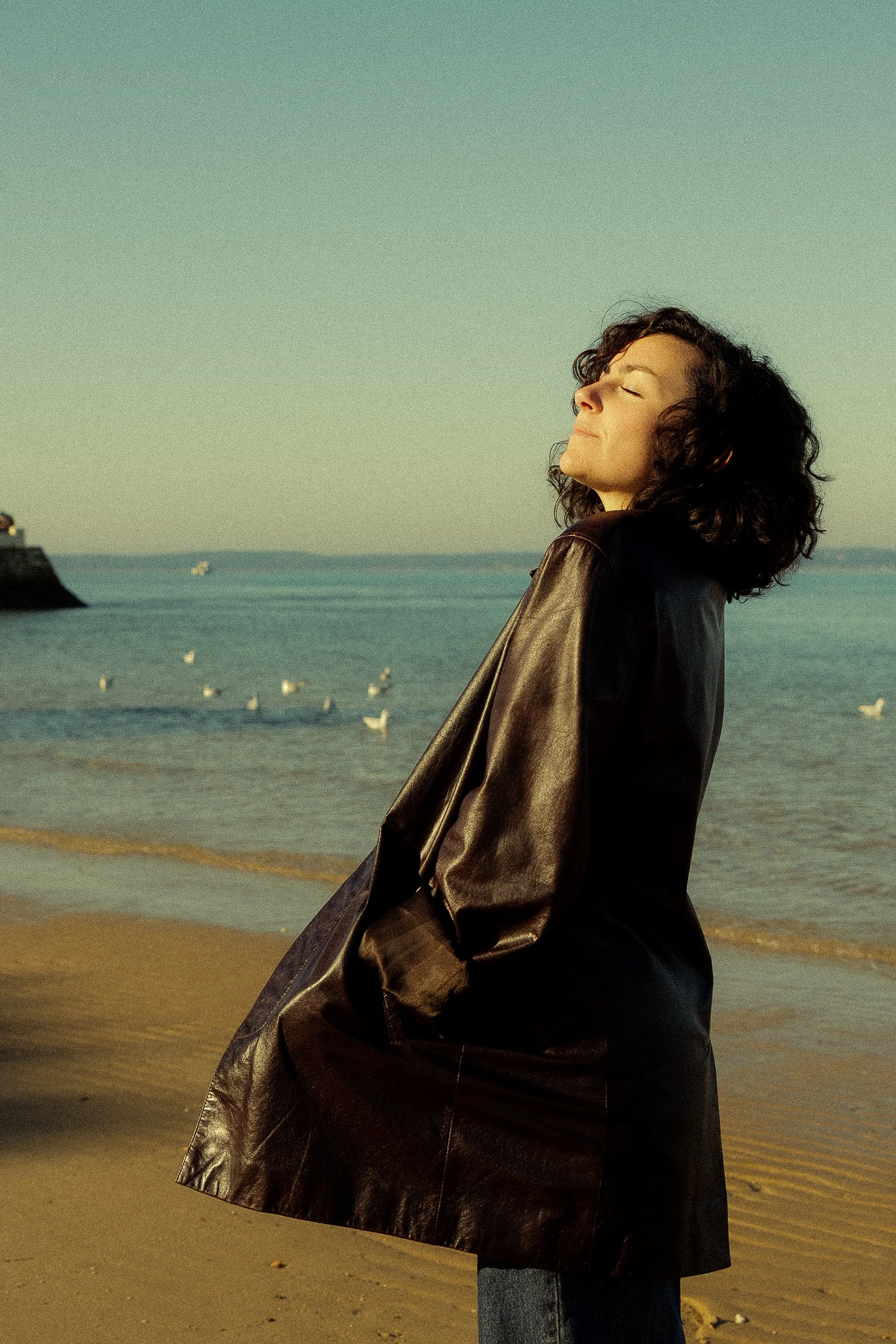 A woman with curly dark hair standing on a beach with her eyes closed and a peaceful expression, wearing a leather jacket.