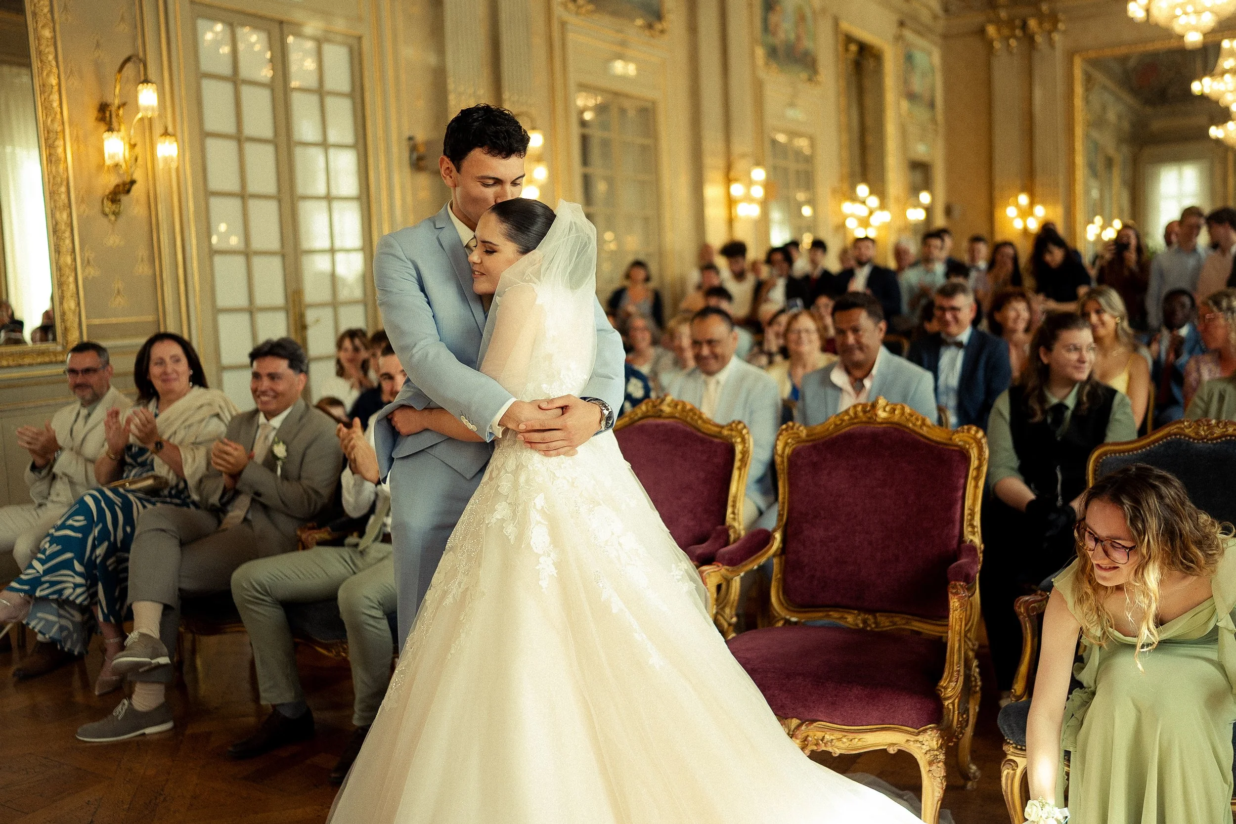 Un couple de mariés en train de s'embrasser lors de leur mariage, dans une salle élégante remplie de invités applaudissant.