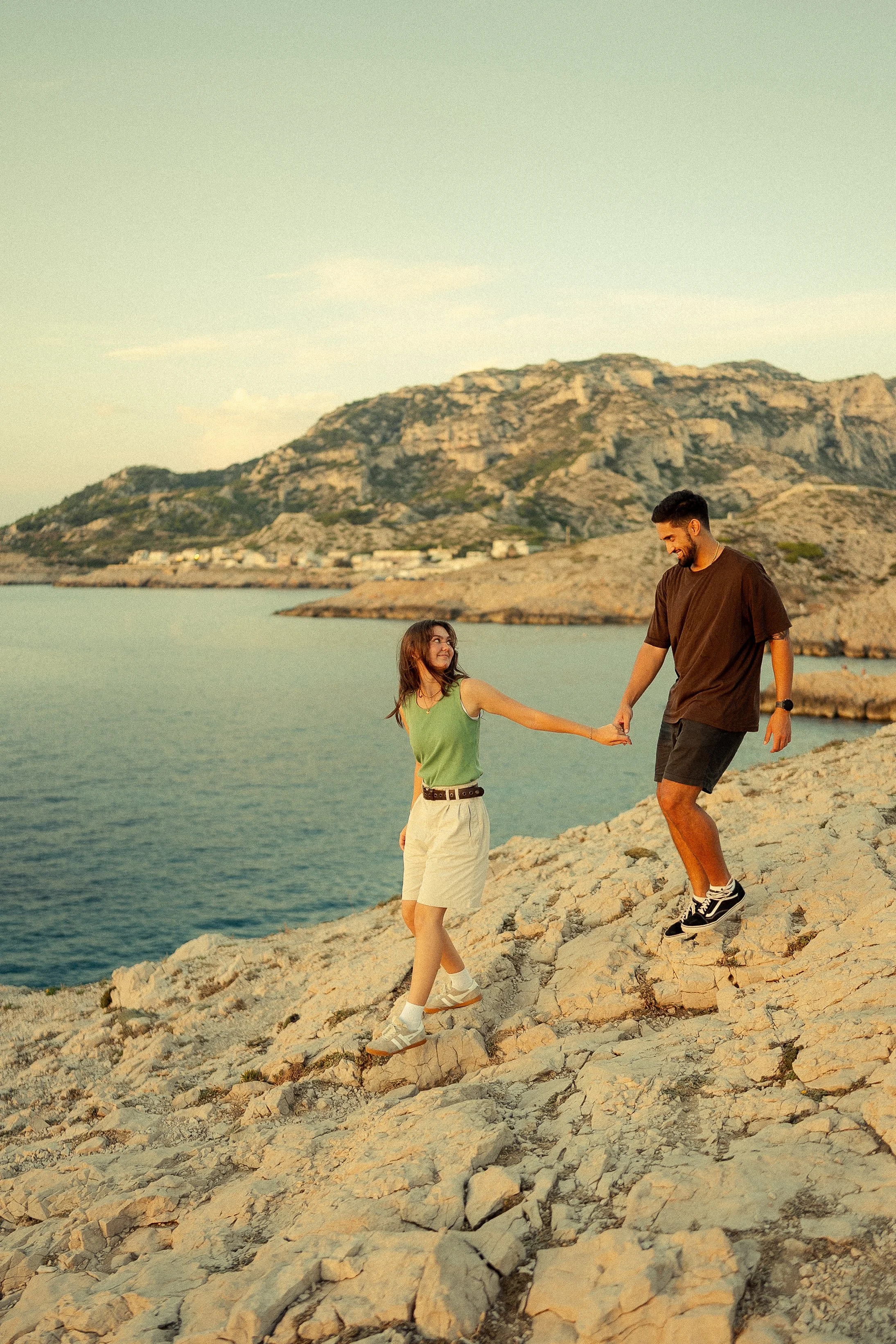 A young couple holding hands and enjoying a walk on rocky terrain near a body of water with mountains in the background during sunset.