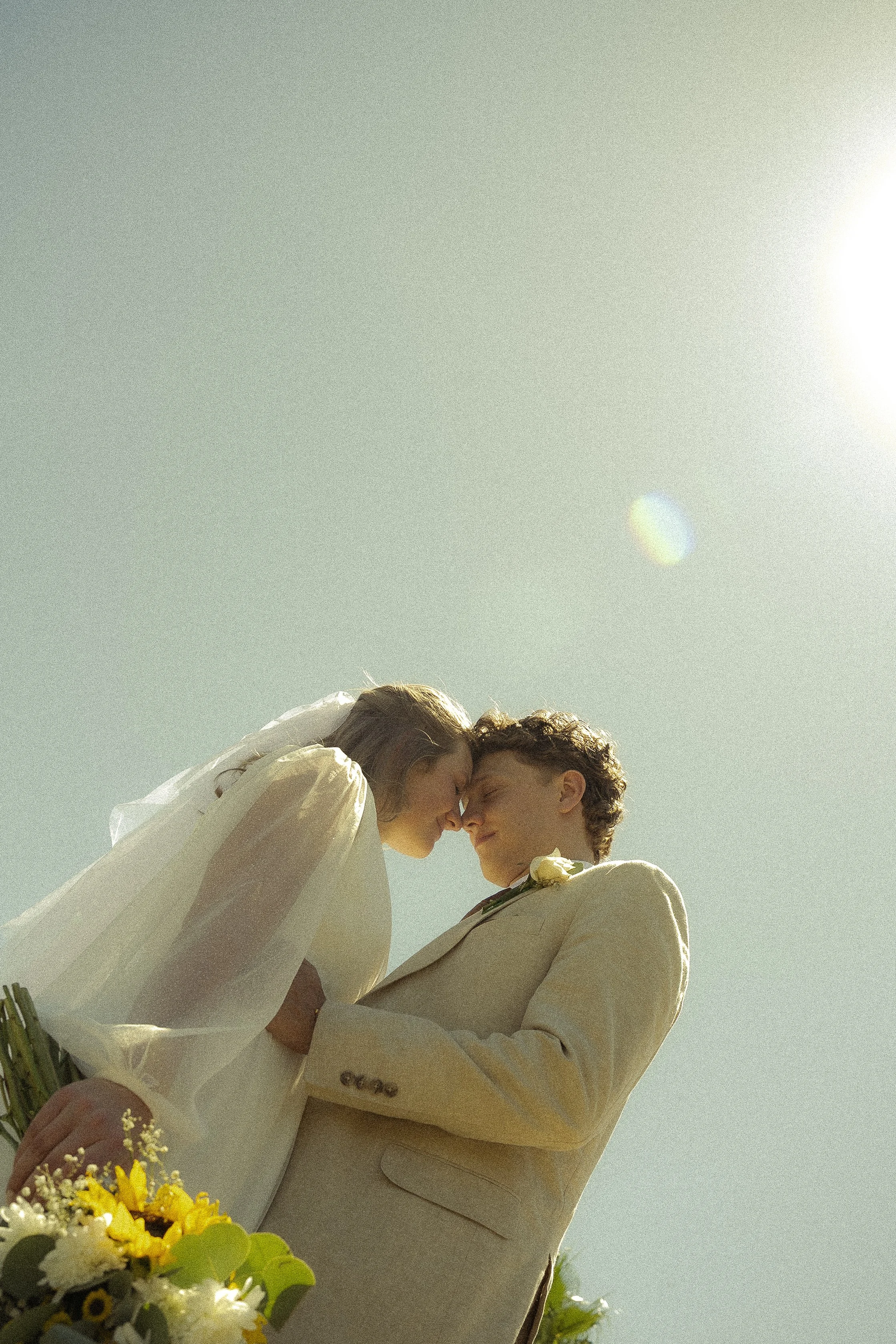 Un couple qui se tient face à face, les yeux fermés, lors d'une cérémonie en plein air, avec un ciel clair en arrière-plan, tenant un bouquet de fleurs dans les mains.