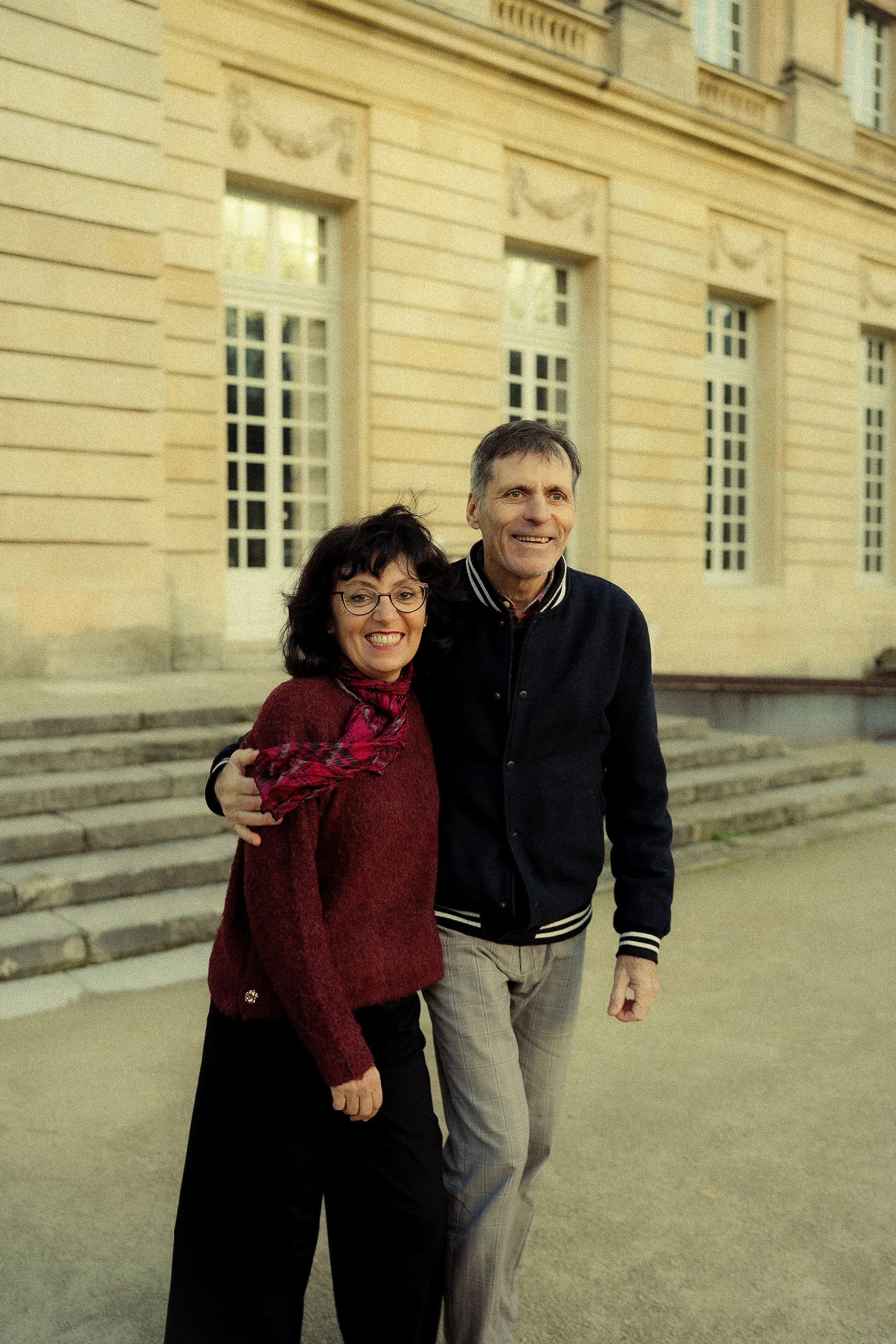 A smiling woman and man embracing outdoors in front of a historic building with stairs, windows, and decorative architectural details.