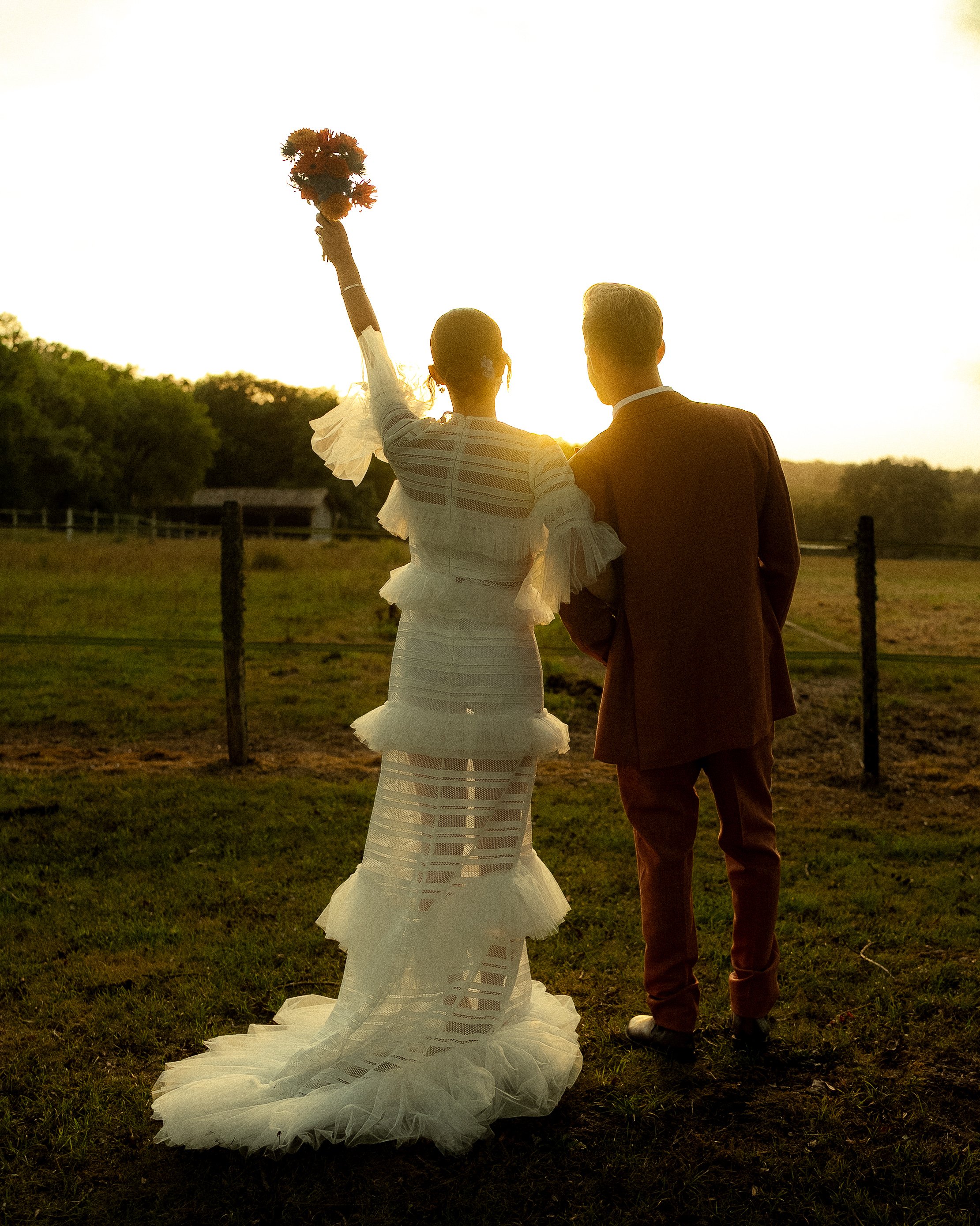A bride and groom standing outdoors on a grassy field at sunset, with the bride holding a bouquet of flowers raised in the air.