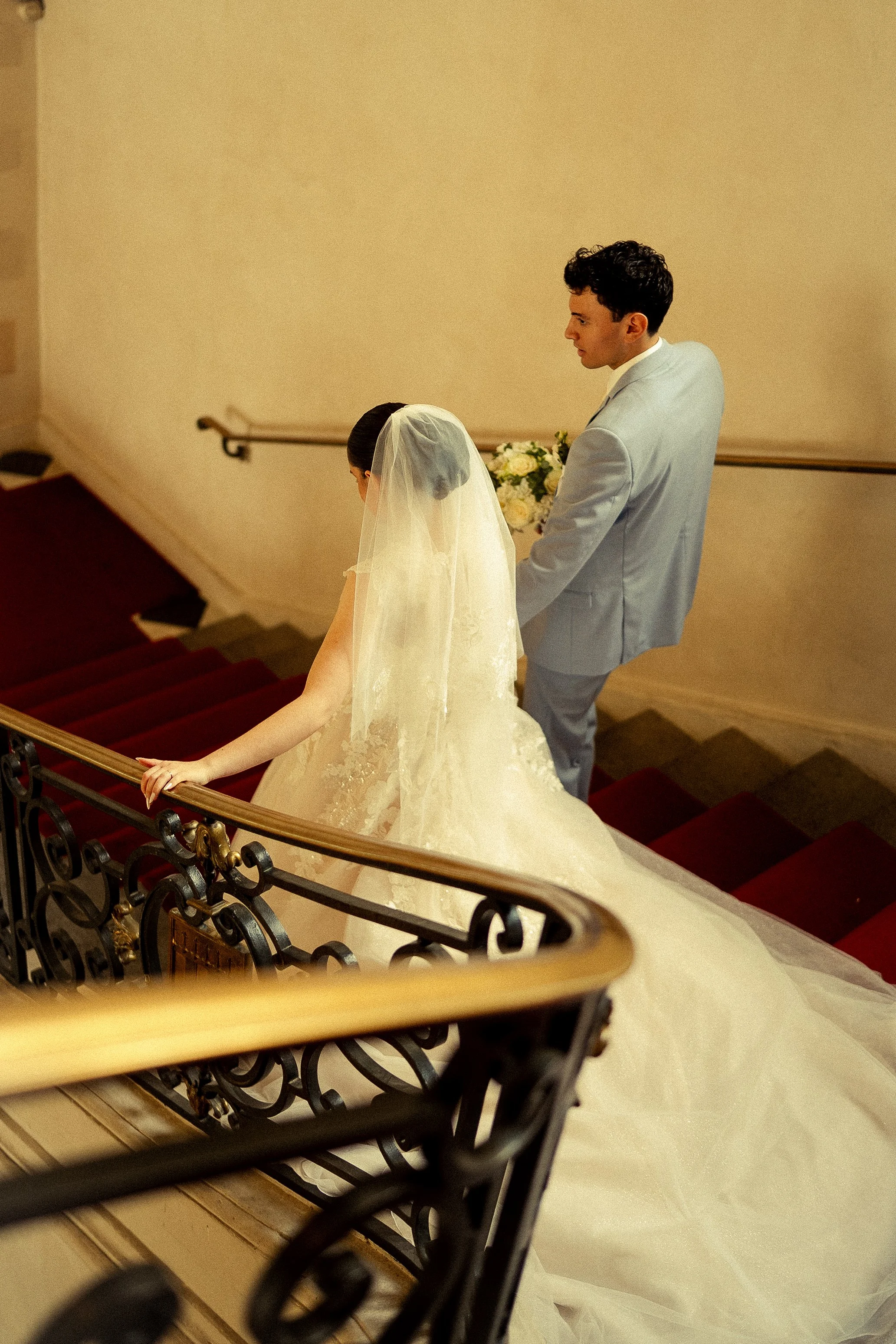 Bride and groom on a staircase during their wedding, with the bride wearing a white wedding gown and veil, and the groom in a light gray suit holding a bouquet of white flowers.