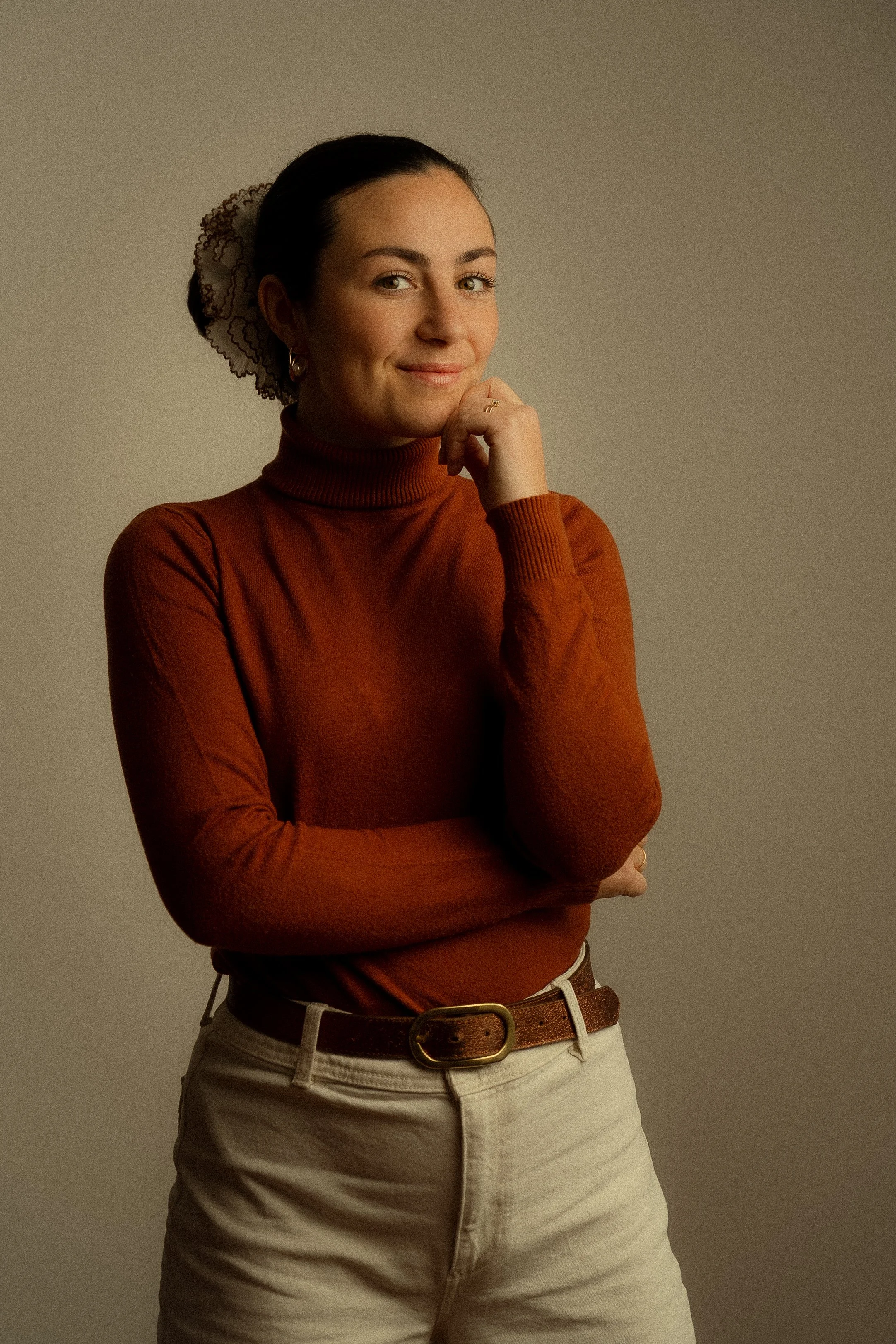 Une femme en haut à col roulé orange, portant un pantalon beige avec une ceinture en cuir brun, sourit en regardant l'appareil photo, en appuyant son menton sur sa main droite.