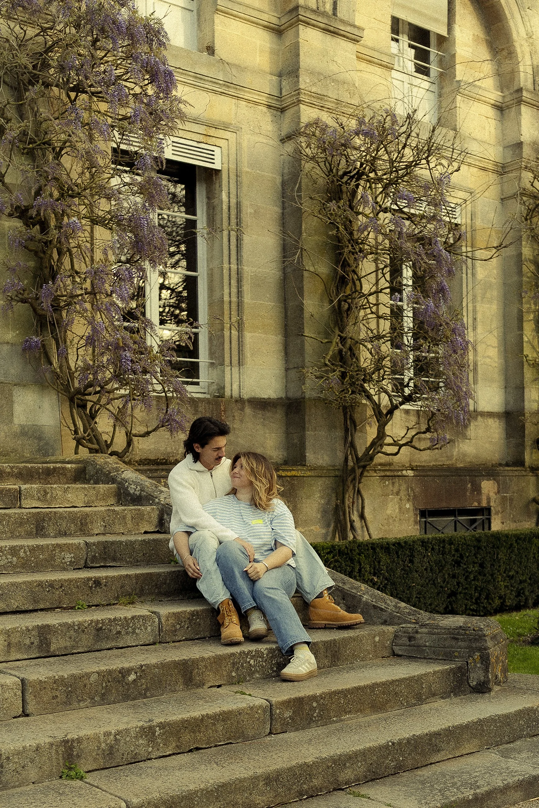 Un homme et une femme assis sur des marches en pierre devant un bâtiment en pierre avec des fenêtres. Les deux personnes semblent discutant ou partageant un moment intime. Il y a deux arbres avec des branches sans feuilles portant des fleurs violettes à l'arrière-plan.