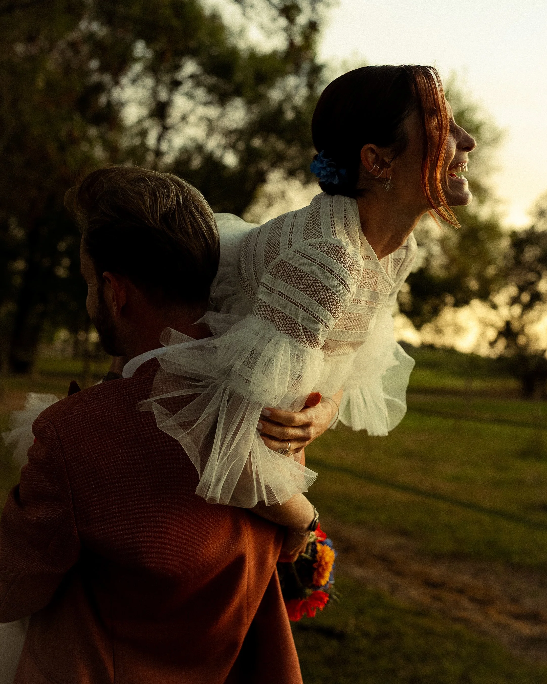 A woman riding on a man's shoulders outdoors at sunset, smiling and laughing. She has short dark hair with a blue flower accessory, wearing a white lace top with puffed sleeves. The man is wearing a brown jacket. The background features trees and a clear sky.