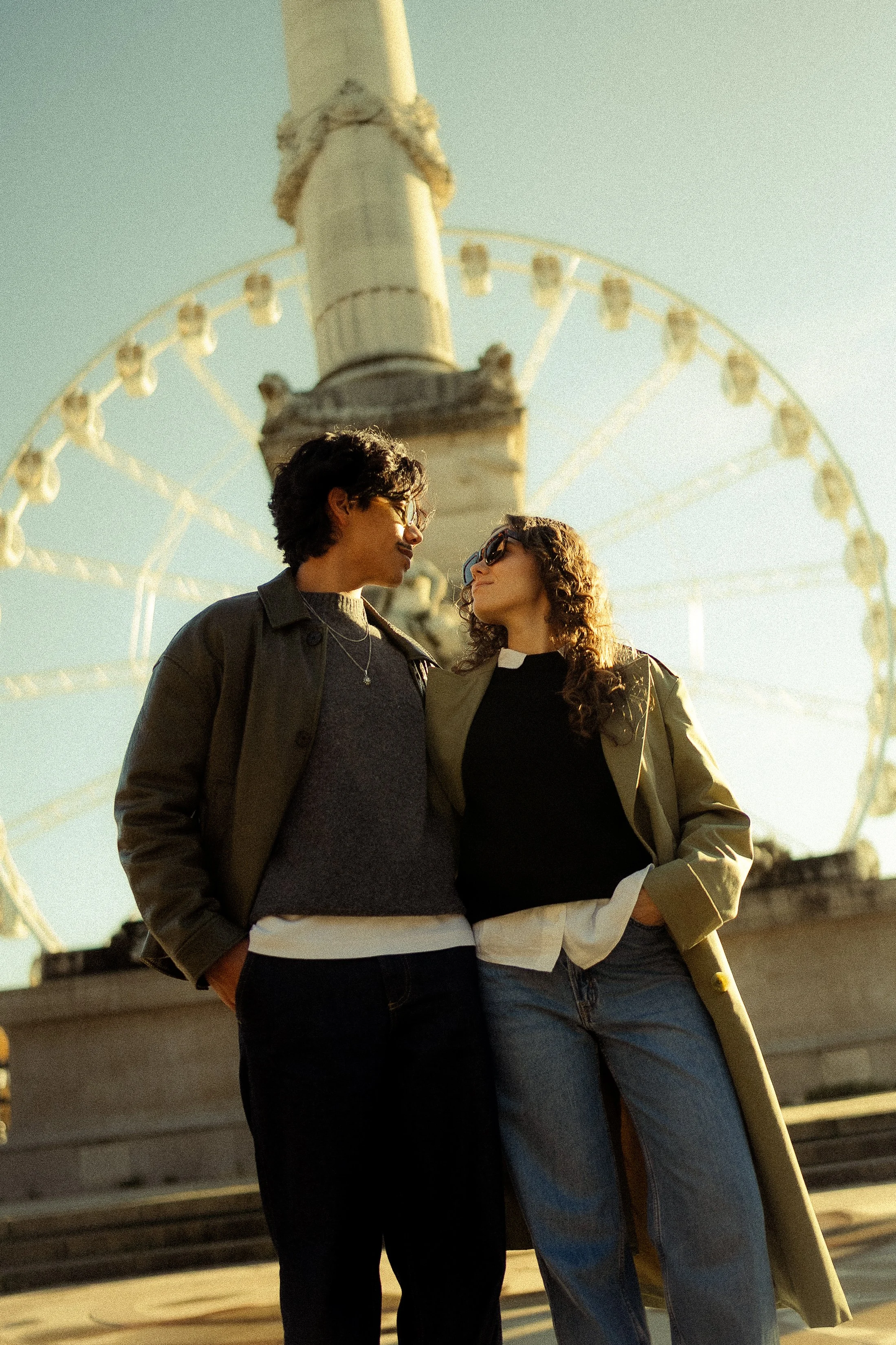 Two women standing close together near a Ferris wheel, facing each other, with the sky in the background.