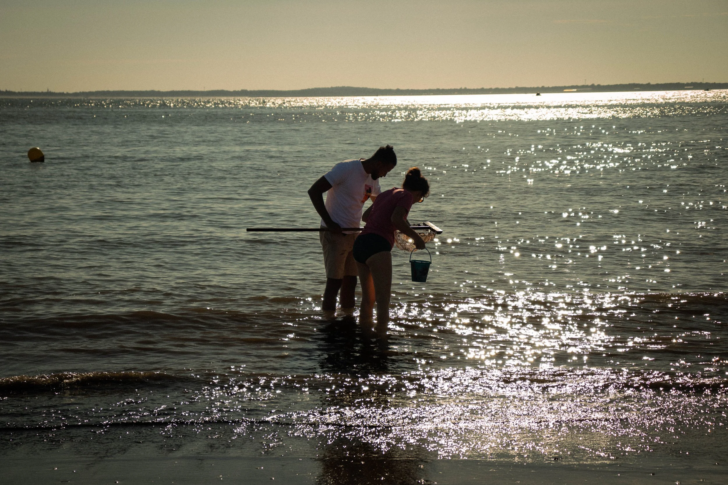 Two people, a man and a woman, standing in shallow water at the beach during sunset, holding a fishing pole, a fishing net, and a bucket.