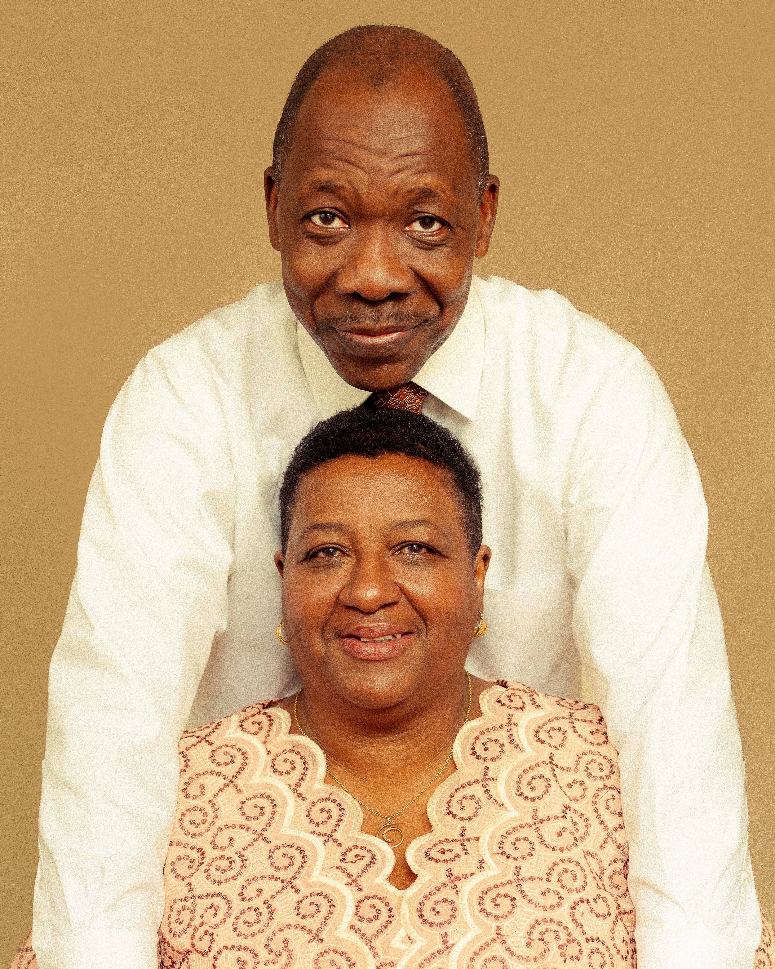 A man and woman pose together, the man standing behind the woman, both smiling and looking at the camera. The man is wearing a white shirt, and the woman is wearing a light-colored, embroidered top with gold jewelry.