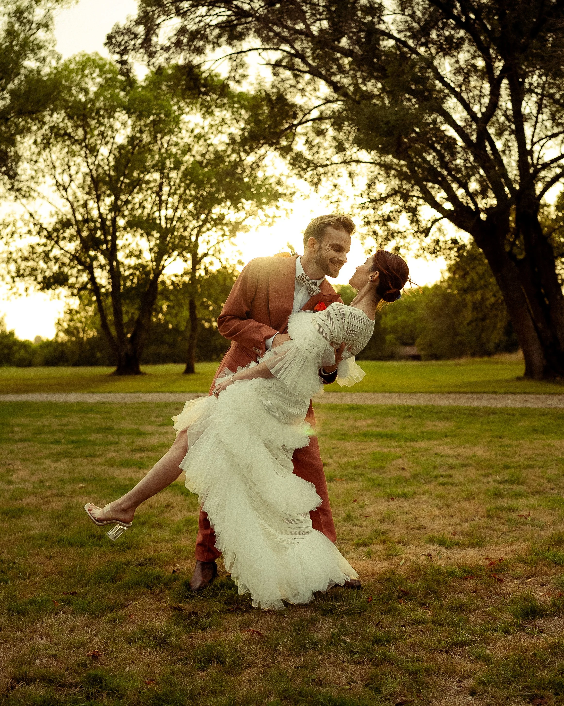 A couple in wedding attire dancing outdoors during sunset, surrounded by trees.