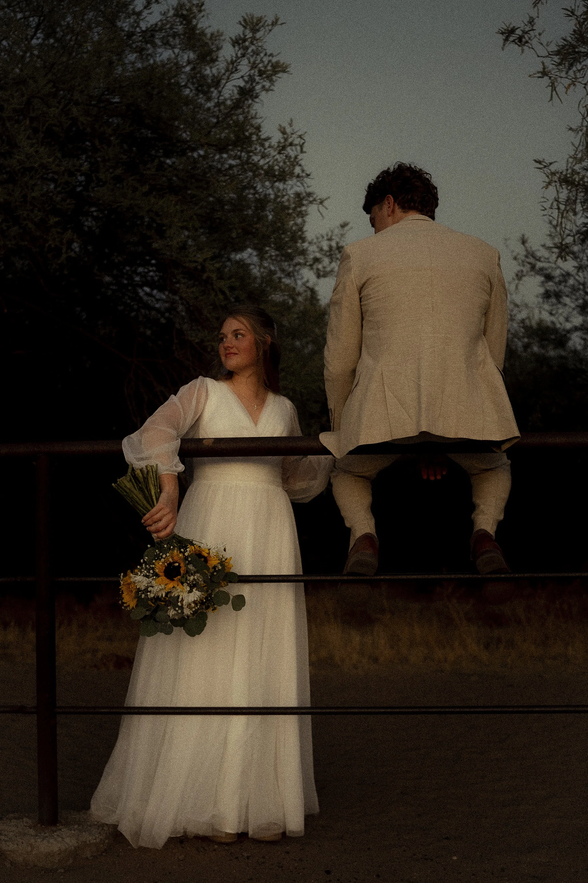 Une femme en robe blanche tient un bouquet de fleurs, un homme en costume beige est assis sur une barrière en bois, en pleine nature au crépuscule.