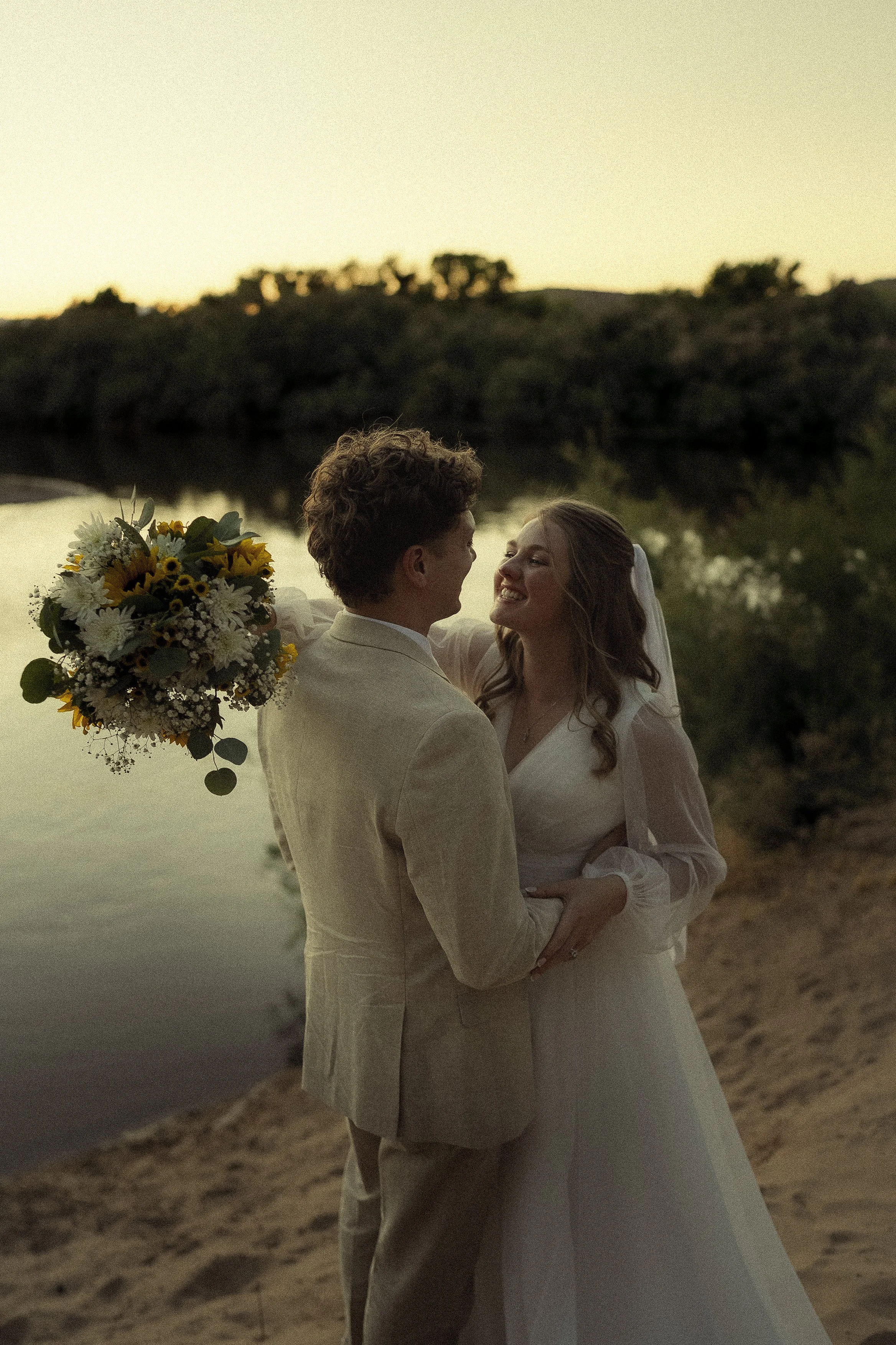 Un couple en mariage se regardant avec bonheur devant un paysage au coucher du soleil, la femme porte une robe blanche et le homme un costume clair, avec un bouquet de fleurs dans la main.