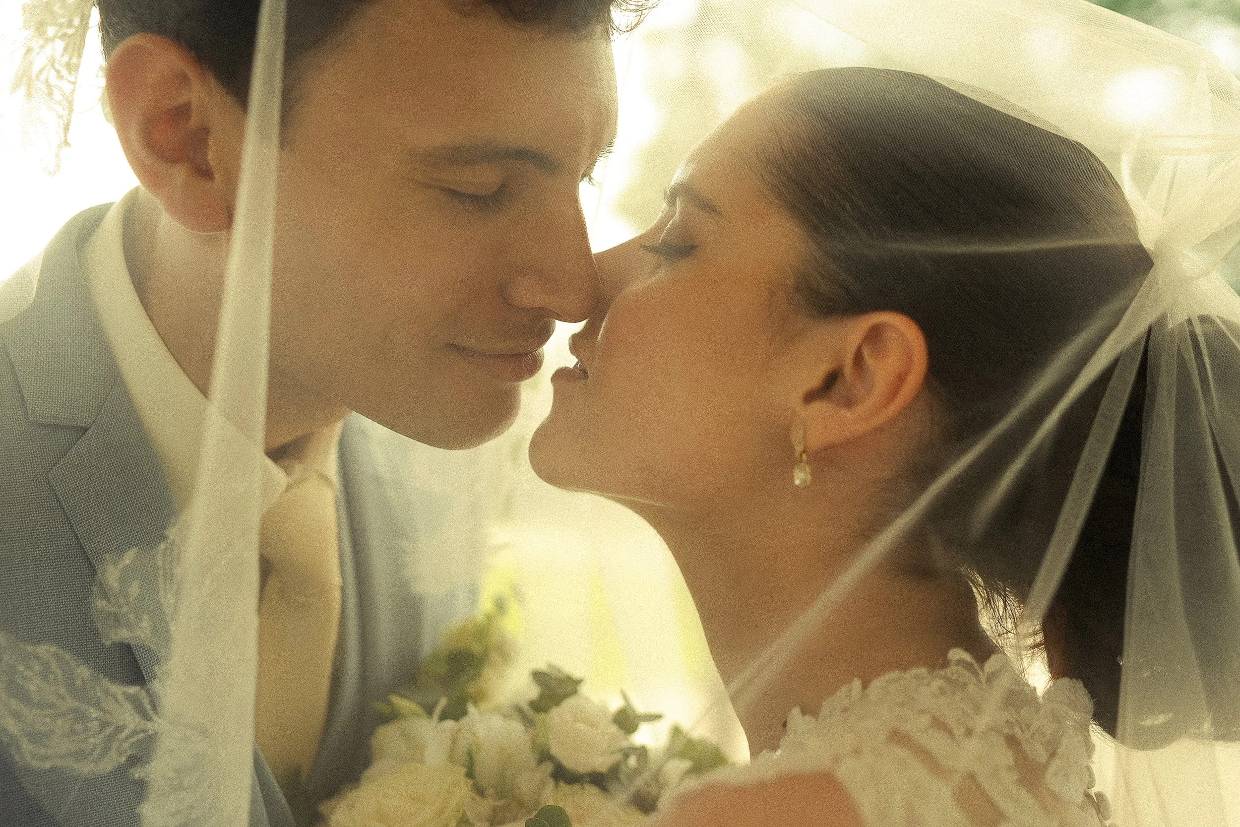 A bride and groom are close together, touching foreheads in a romantic pose, with a soft, warm glow around them.
