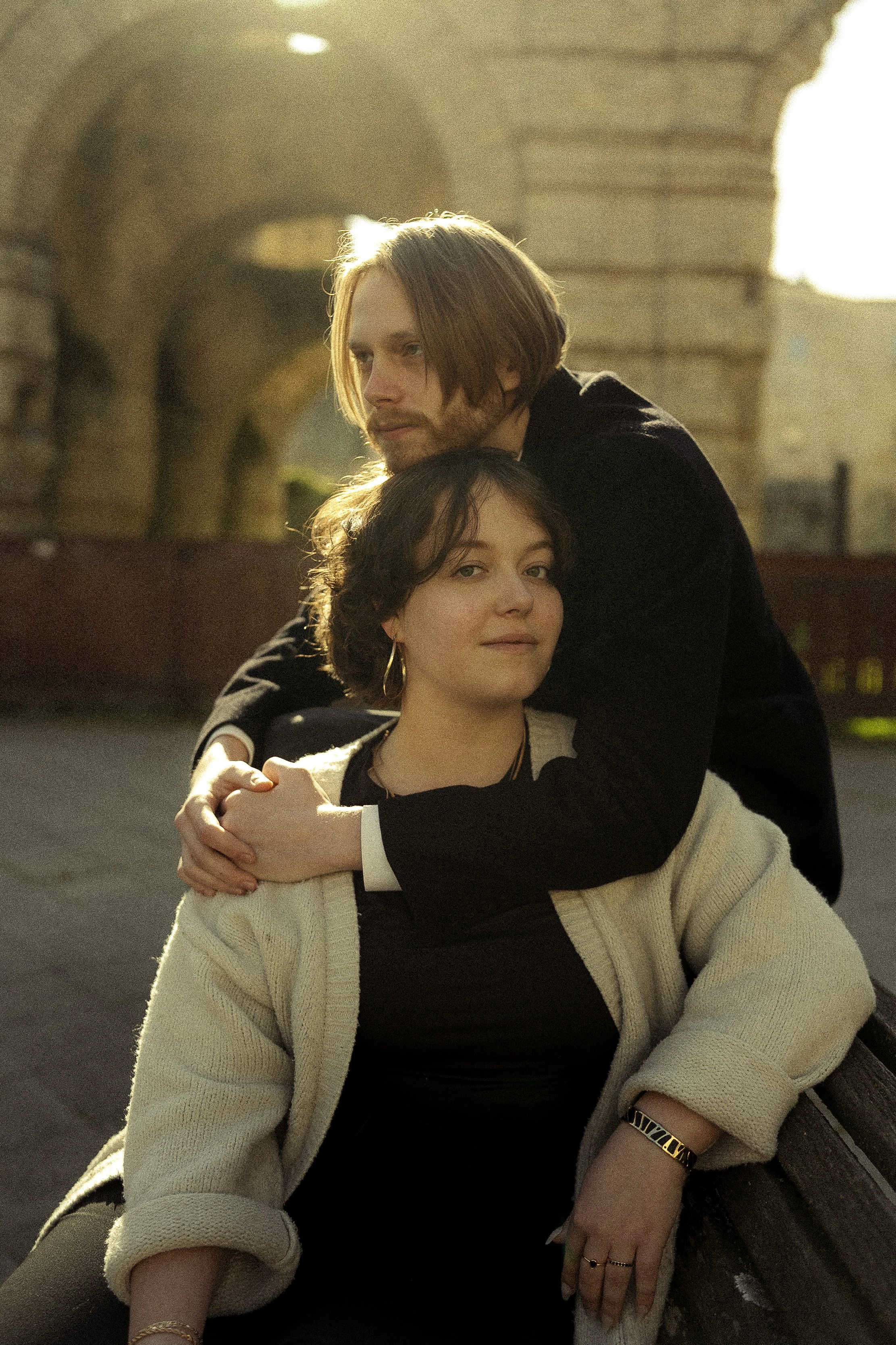 Une femme en pull blanc et un homme en noir sont assis ensemble, l'homme repose sa tête sur la femme. Ils sont sous un arc en pierre, avec la lumière naturelle du soleil derrière eux, créant une atmosphère chaleureuse.