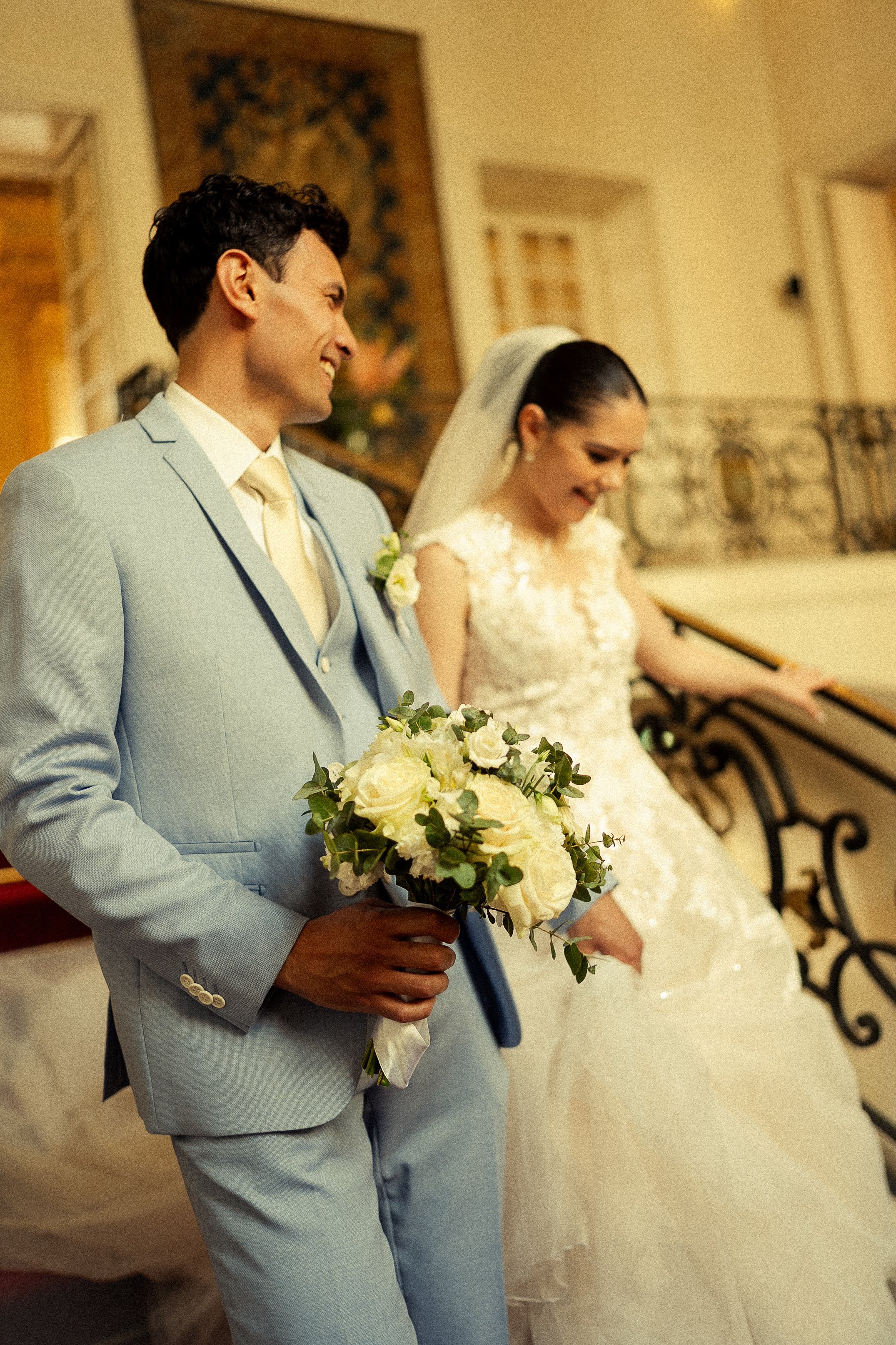 A groom in a light blue suit holding a bouquet of white roses and greenery, standing next to a smiling bride in a white lace wedding dress.