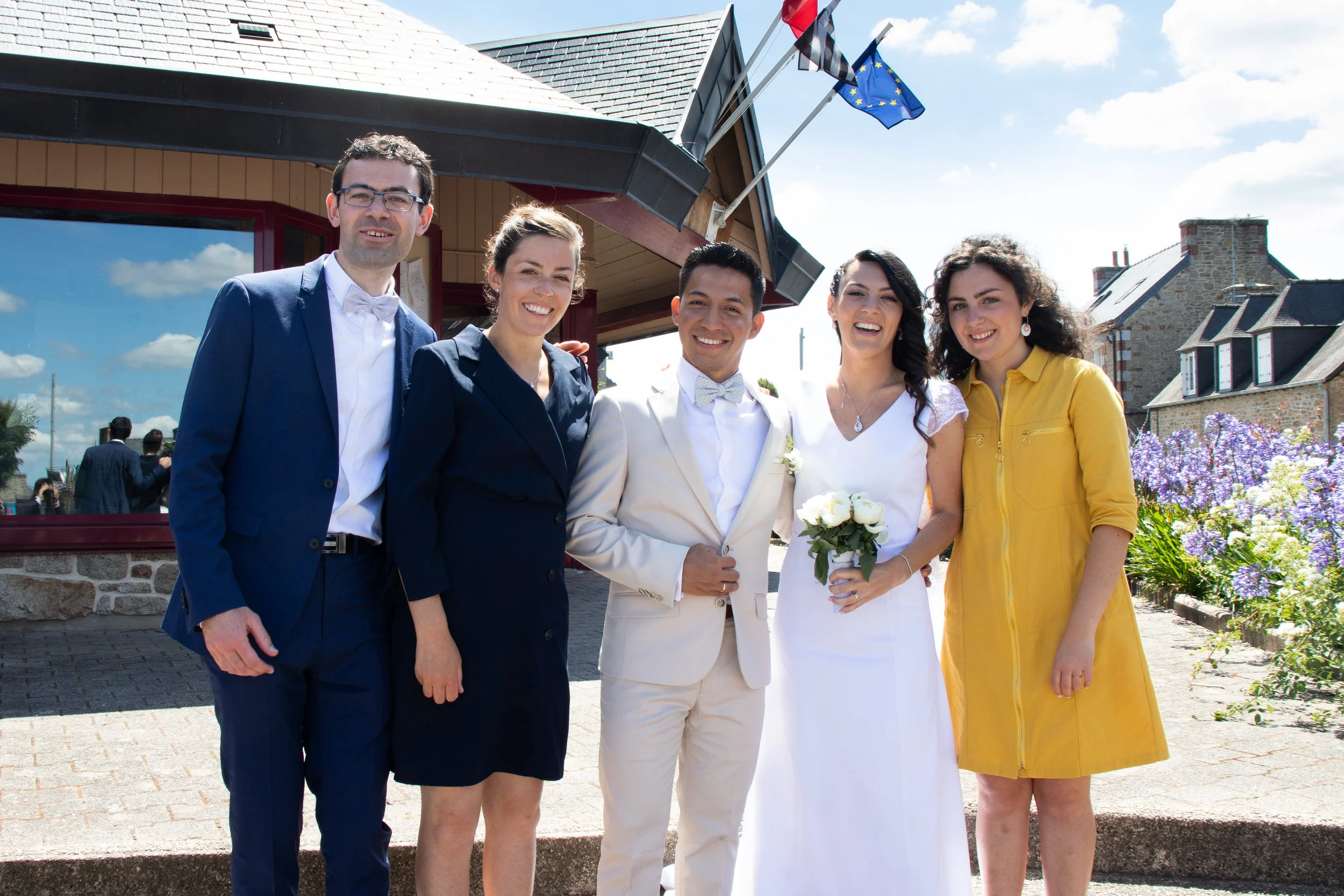 A group of five people at a wedding outdoors, with a building and flowers in the background. The bride is in a white dress holding a bouquet of white flowers, and the groom is next to her in a white suit and bow tie. Three other women and one man are with them, all smiling.