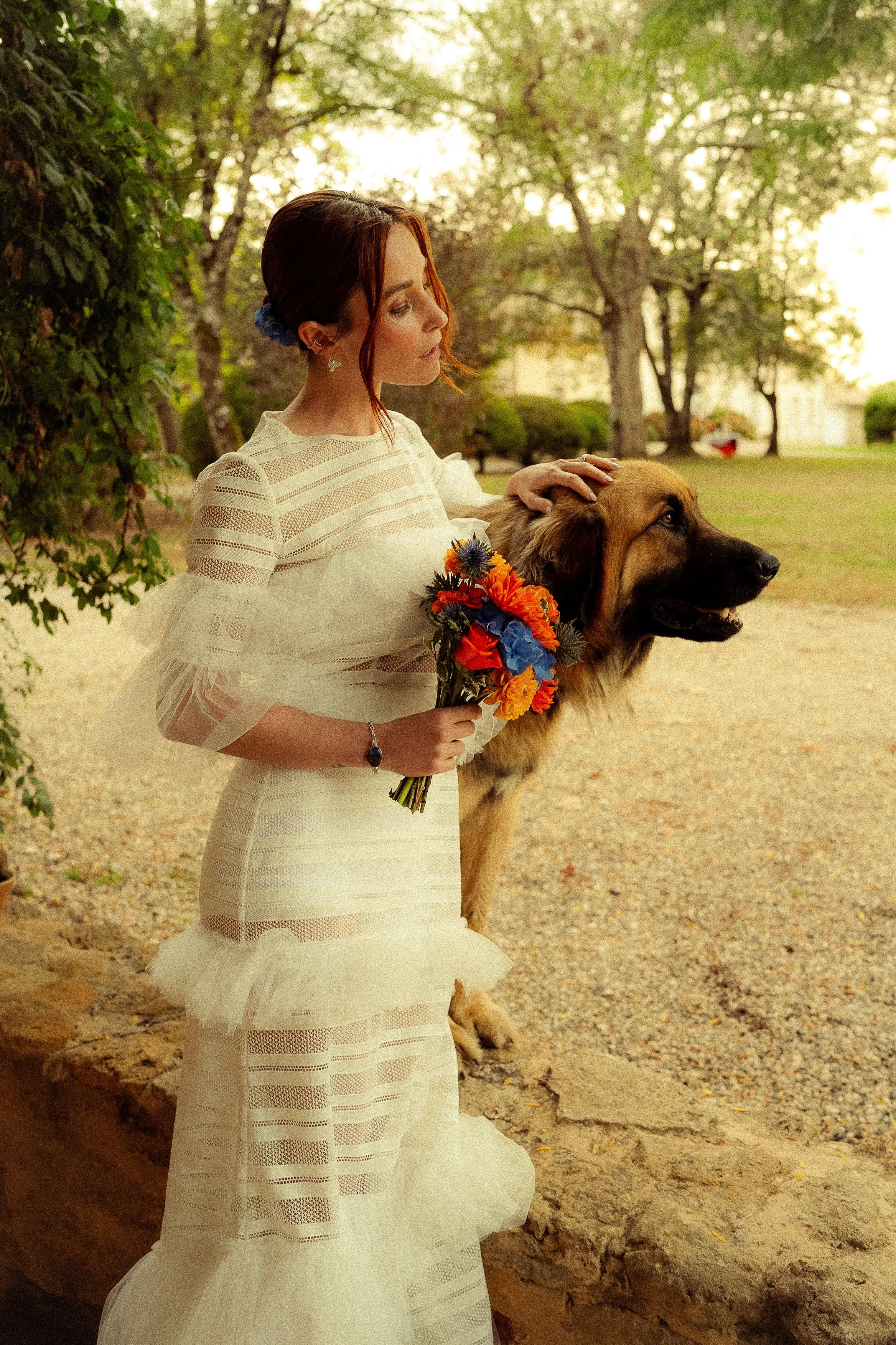 Une femme en robe de mariage blanche avec des détails en dentelle, tenant un bouquet de fleurs colorées, et posant près d'un chien berger allemand dans un parc vert en arrière-plan.