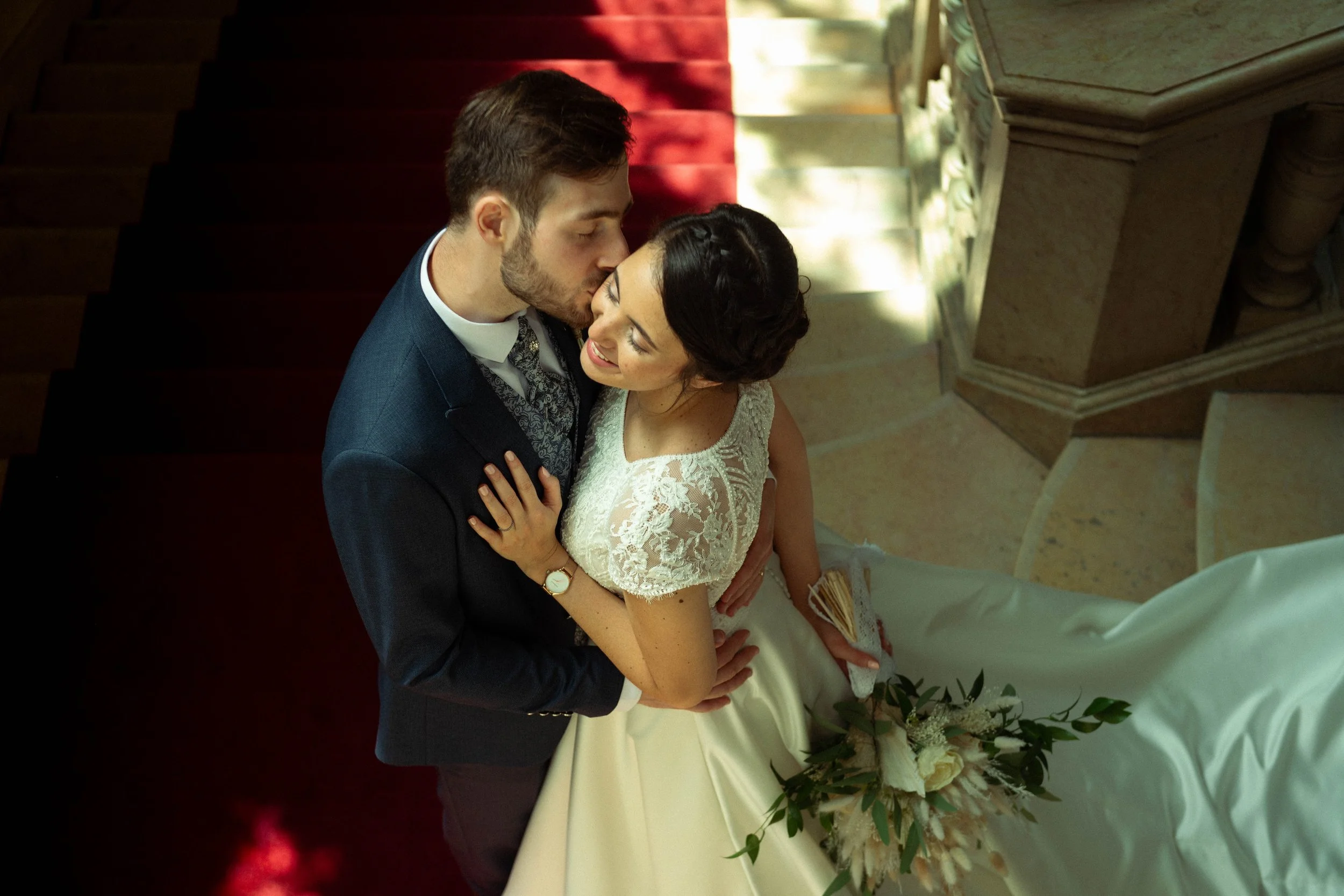 A newlywed couple sharing a kiss on a staircase, with the bride holding a bouquet of white and green flowers and wearing a lace wedding dress, while the groom is dressed in a dark suit with a patterned vest and tie.