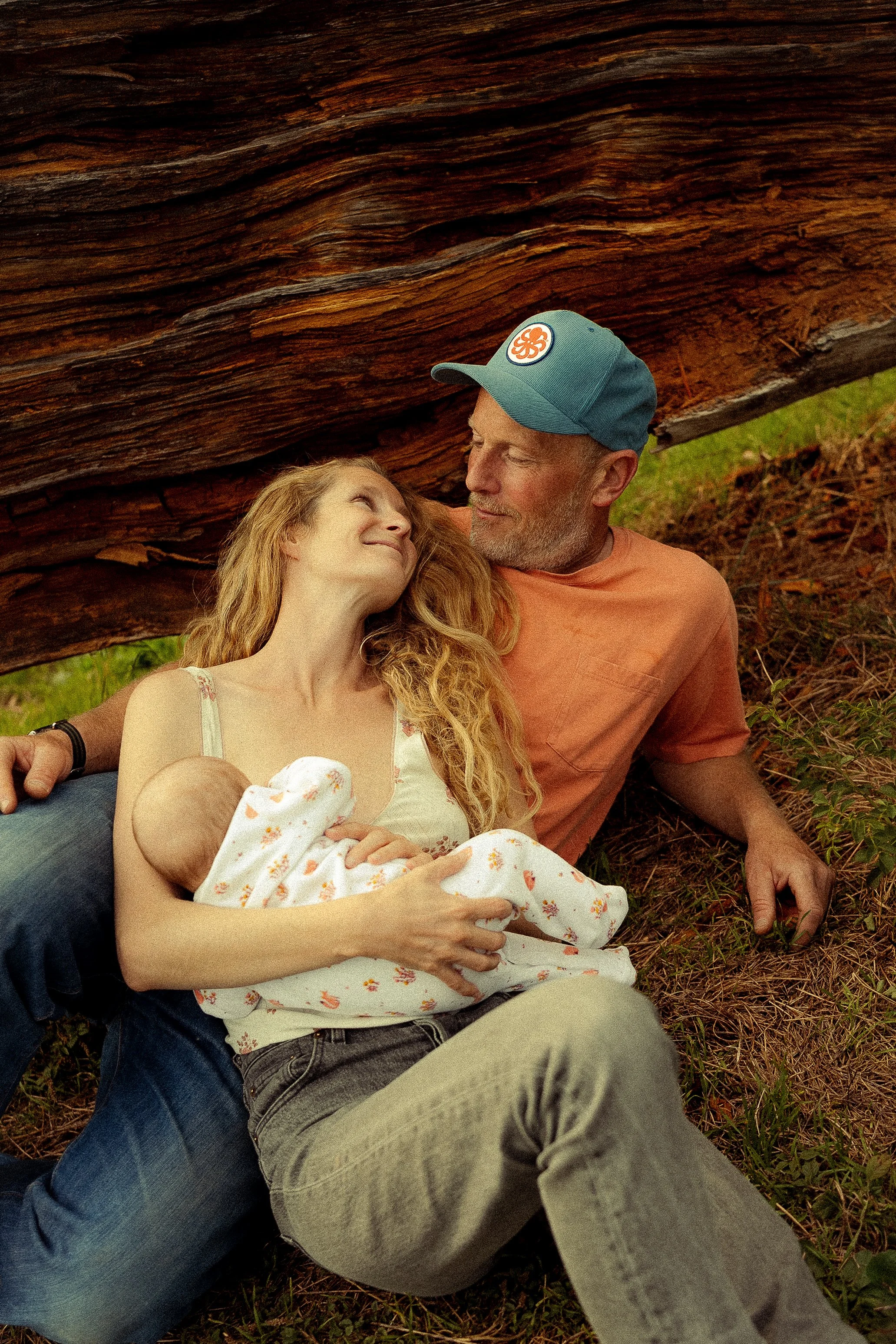 A couple, a woman and a man, sitting on the grass under a large fallen tree with a newborn baby wrapped in a white blanket with orange flowers. They appear to be enjoying a peaceful moment together outdoors.