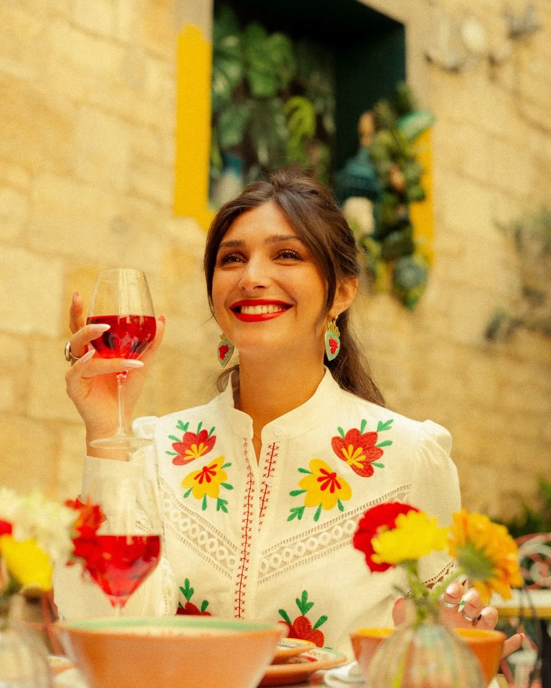 A woman with dark hair and bright red lipstick, smiling and holding a glass of red wine, sitting at a table decorated with colorful flowers, wearing a white blouse with vibrant embroidered floral patterns.