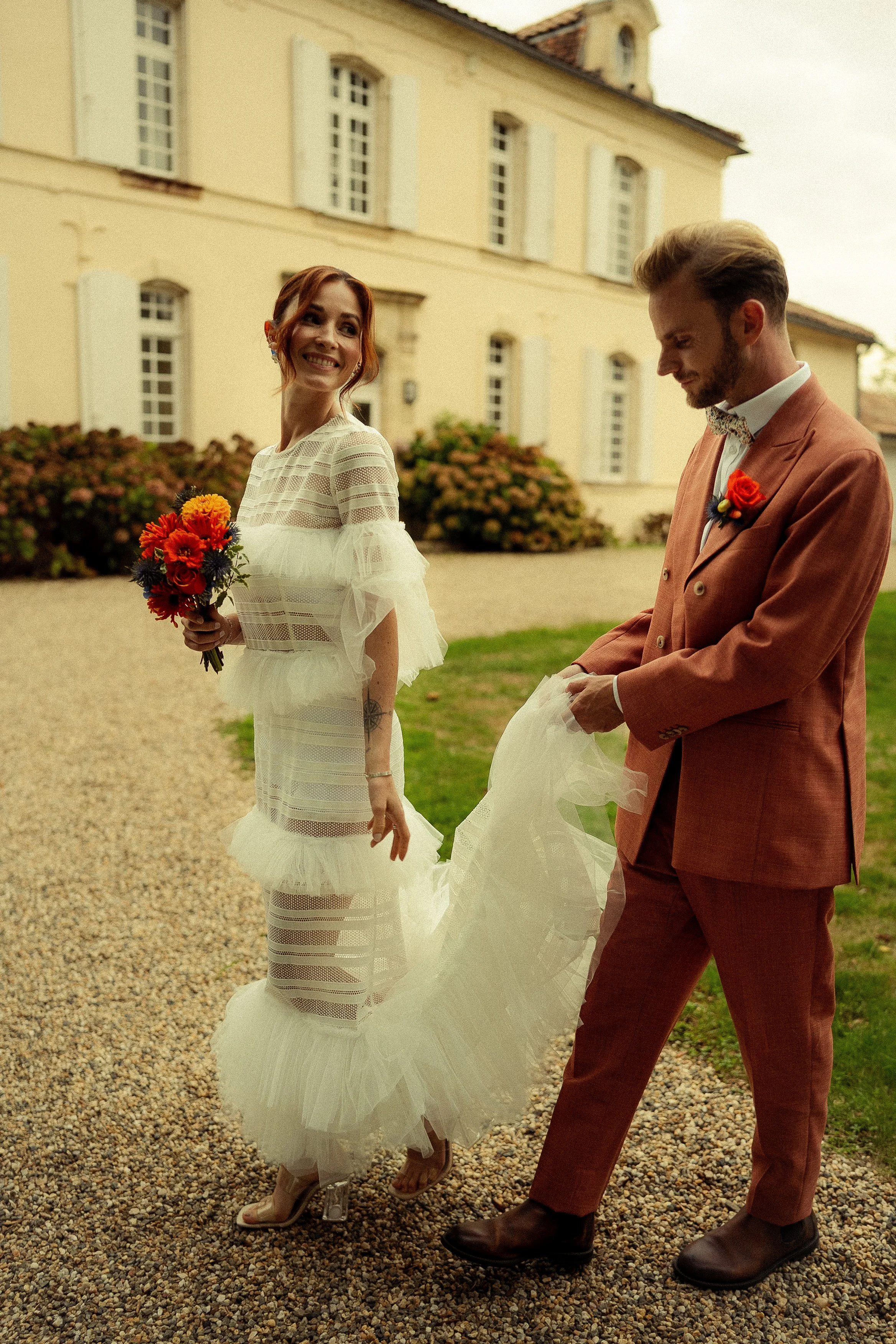 A bride with red hair smiling and holding a bouquet of orange and purple flowers, standing outdoors next to a groom in a brown suit, near a yellow building with white shutters.