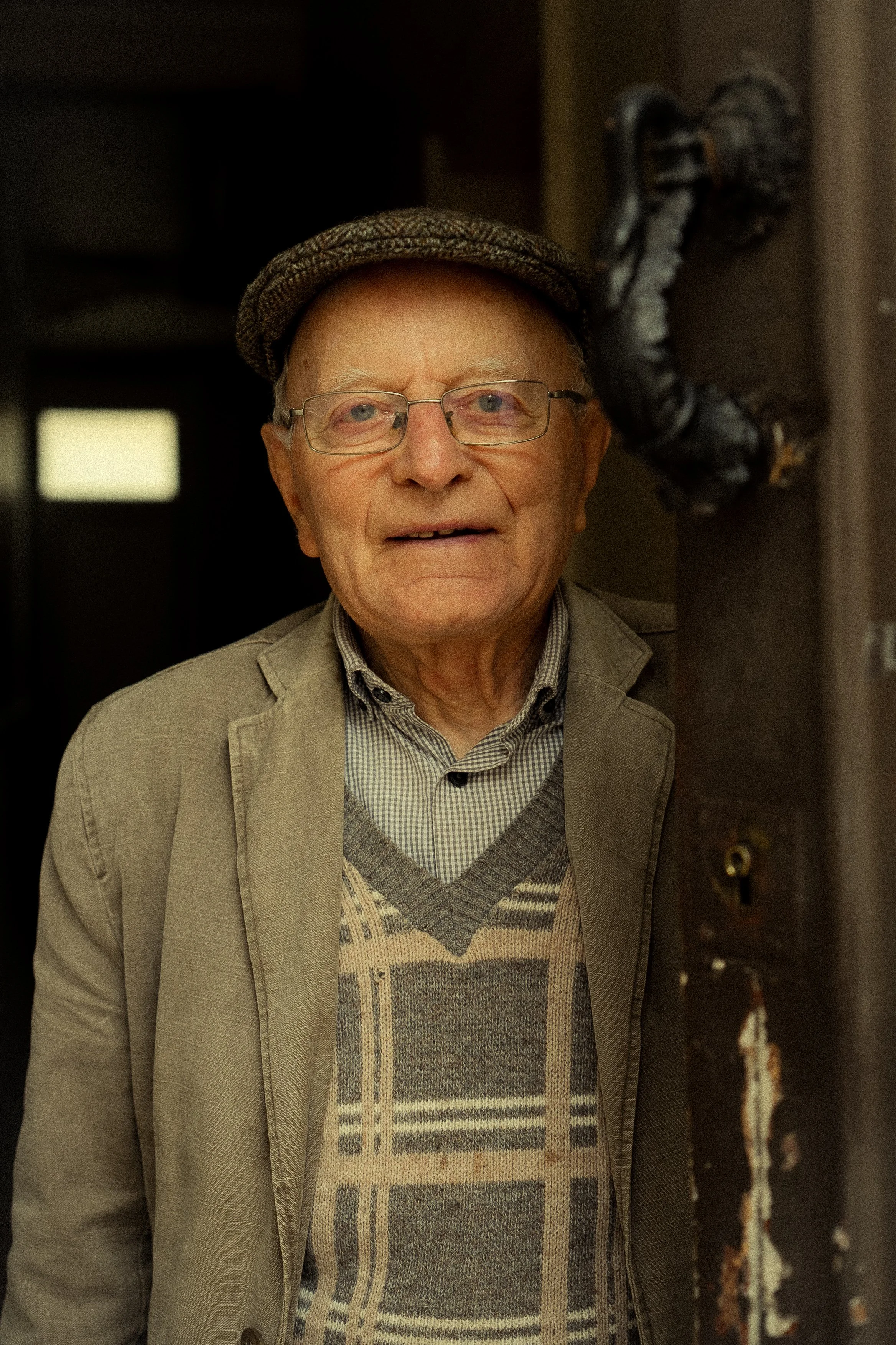 Un homme âgé portant des lunettes, un bonnet et un veston, sourit en regardant l'appareil photo, debout près d'une porte.