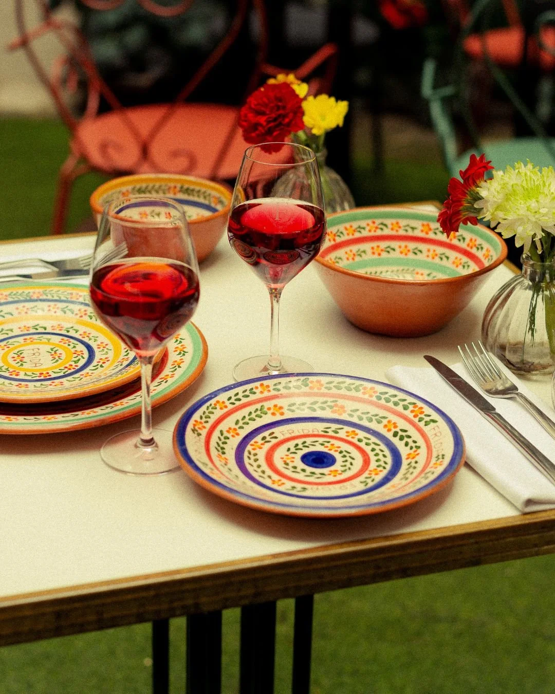 A table set for a meal with colorful patterned plates, two glasses of red wine, and small vases with red, yellow, and white flowers. Background includes chairs and greenery.
