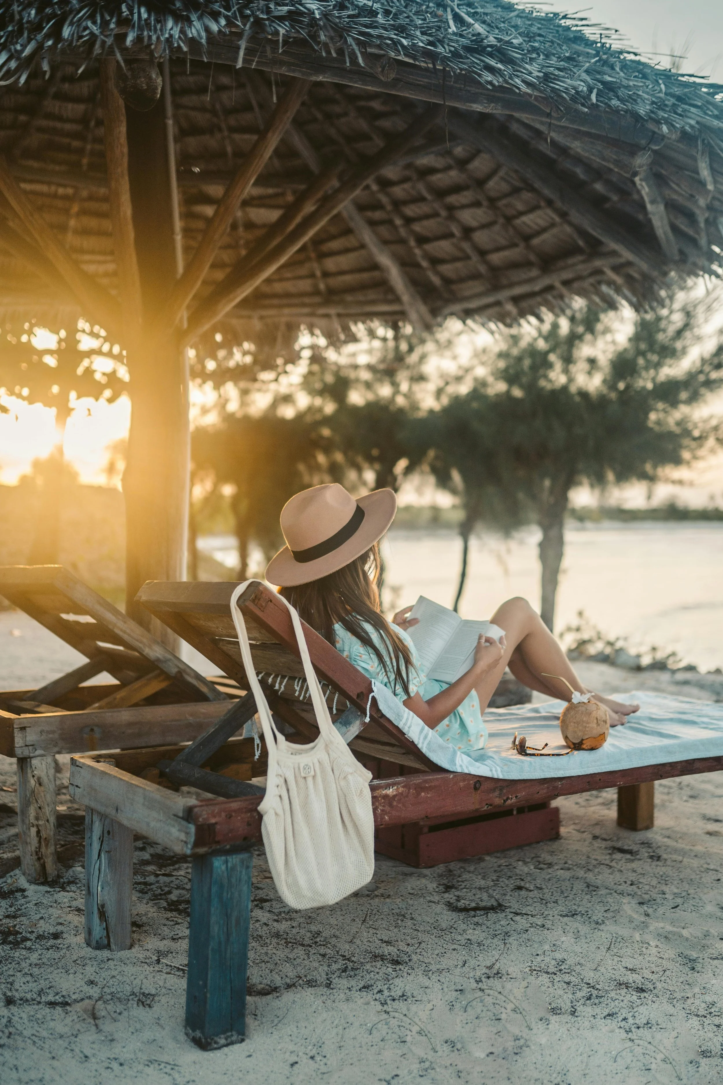 A woman relaxing on a wooden lounge chair under a thatched umbrella on a beach at sunset, reading a book, with a coconut drink nearby, and trees and water in the background.