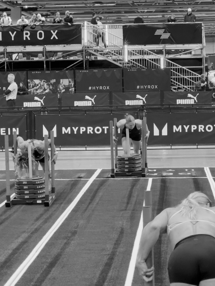 Two women participate in a competitive indoor weightlifting event, lifting weighted sleds, with spectators watching from behind barriers and tiered seating.