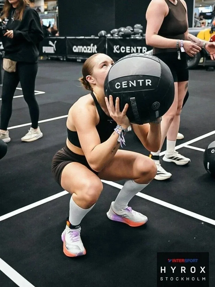A woman in athletic wear is squatting and holding a large black medicine ball labeled 'CENTR' at a gym. She is looking upward and surrounded by other people in workout attire.