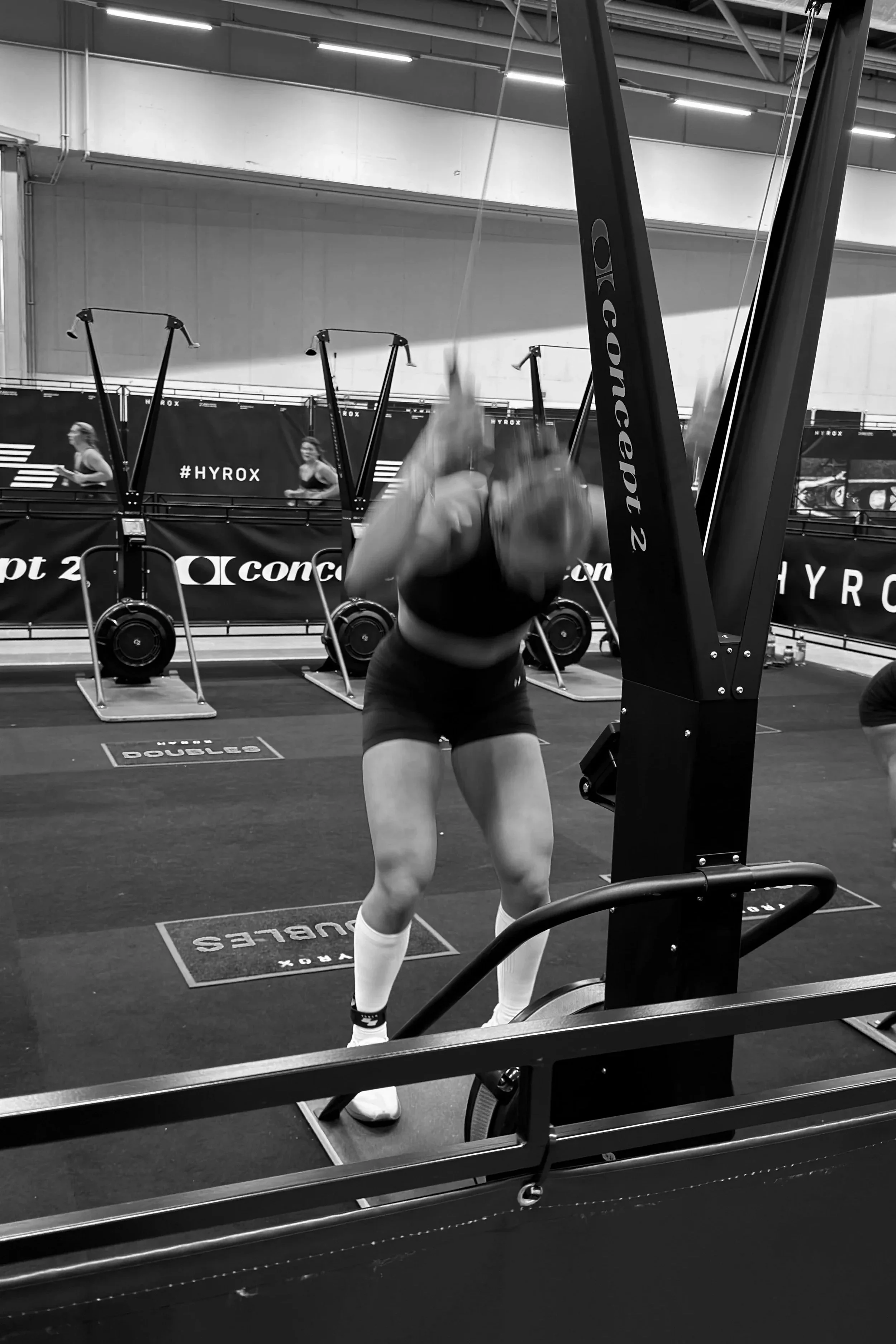 A woman exercising with battle ropes in a gym, with other gym equipment and people in the background.