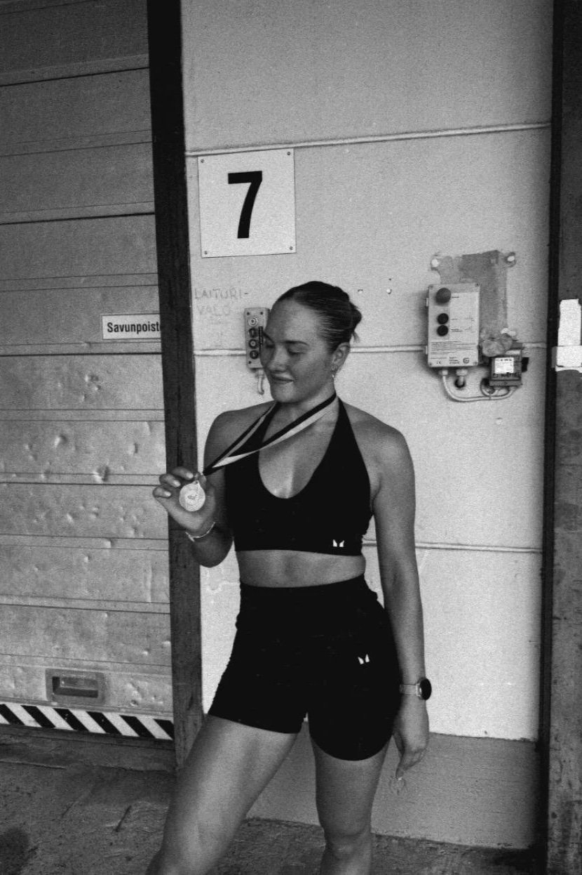 A young woman in athletic attire holding a medal inside a building, smiling, with a large number 7 sign on the wall behind her.