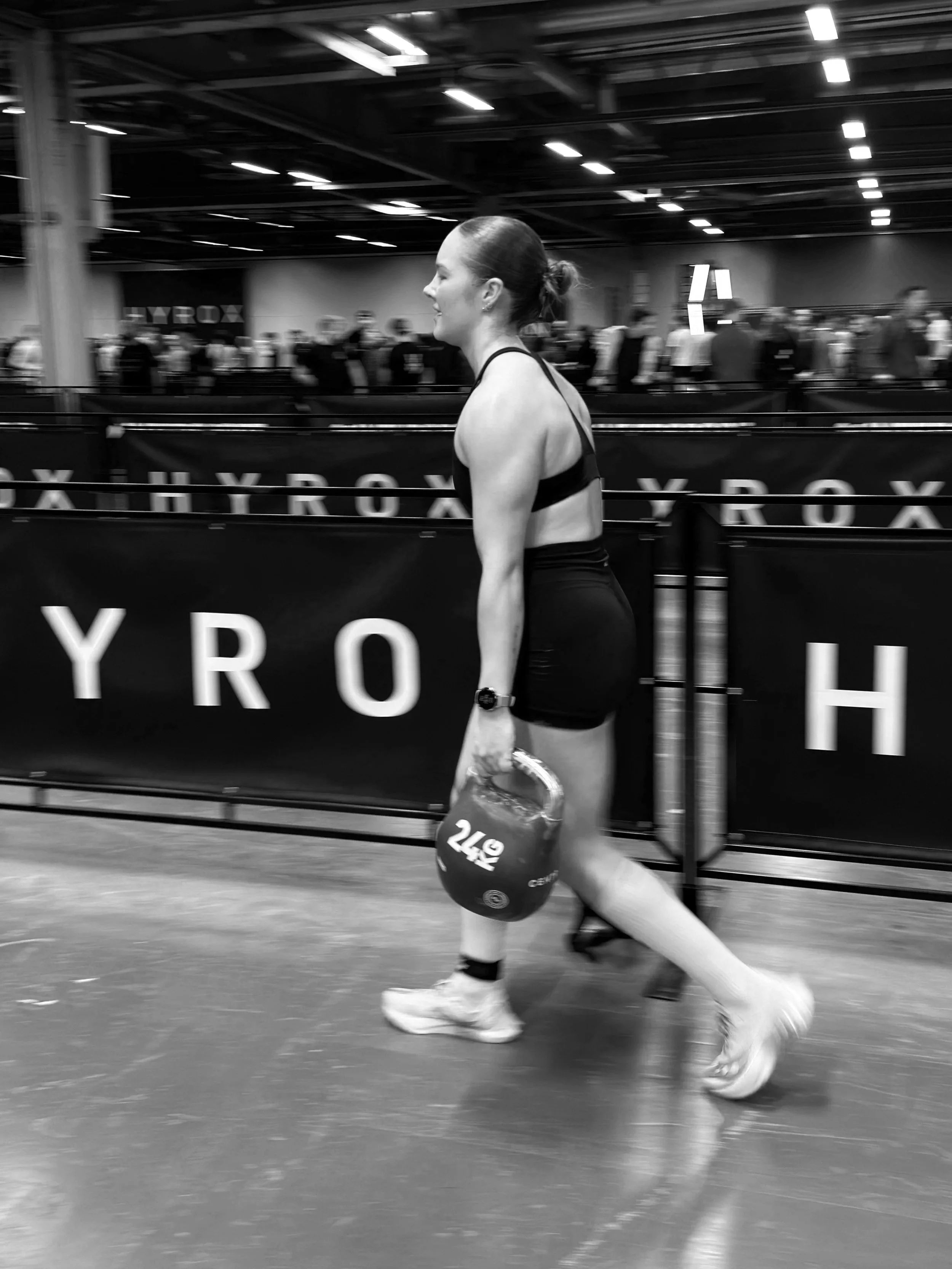 A woman in athletic wear walking inside a gym or sports arena, carrying a helmet. She is surrounded by other people and banners.