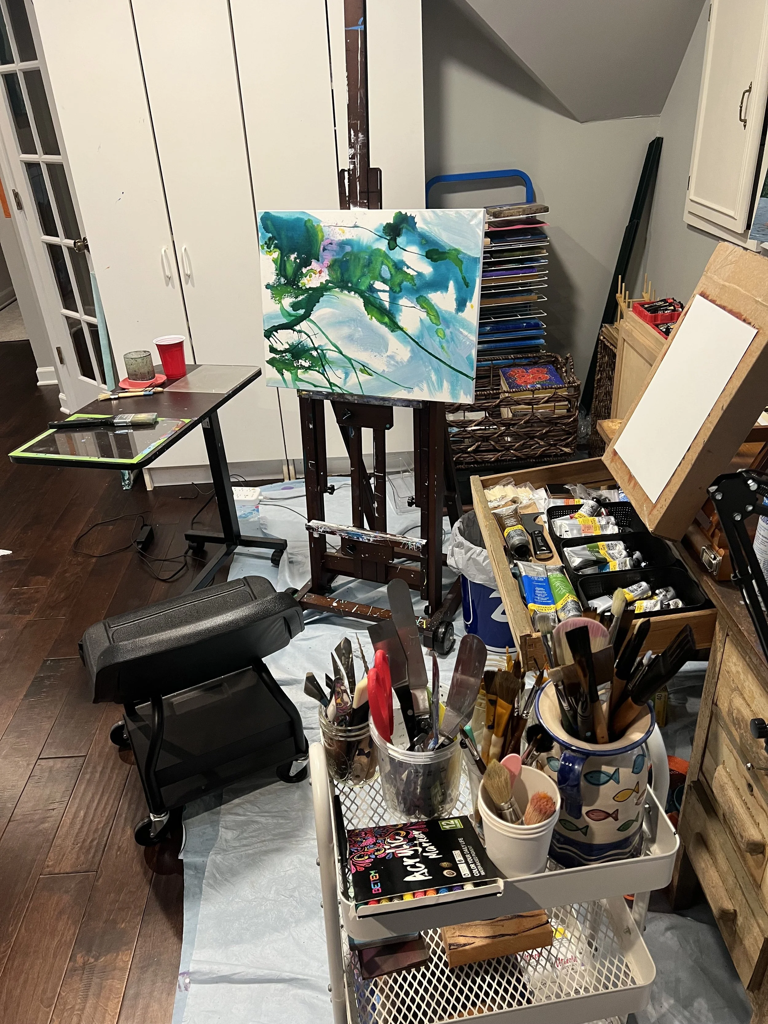 An art studio with a painting of an abstract landscape on an easel, surrounded by art supplies, brushes, paint tubes, and a workspace.