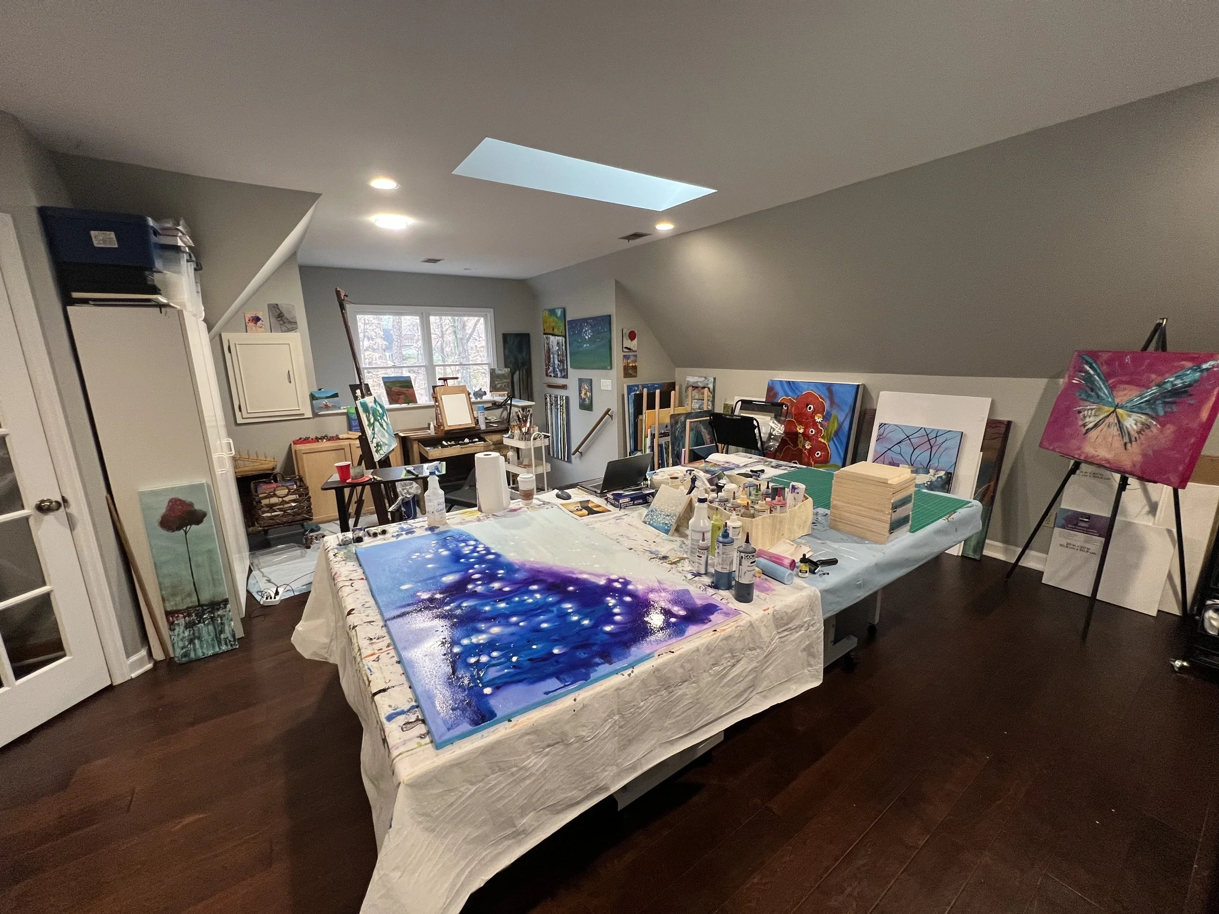Art studio with paintings and supplies on a table, featuring a purple and blue galaxy-themed artwork in progress, various paints, brushes, and canvases in a cozy, well-lit room.