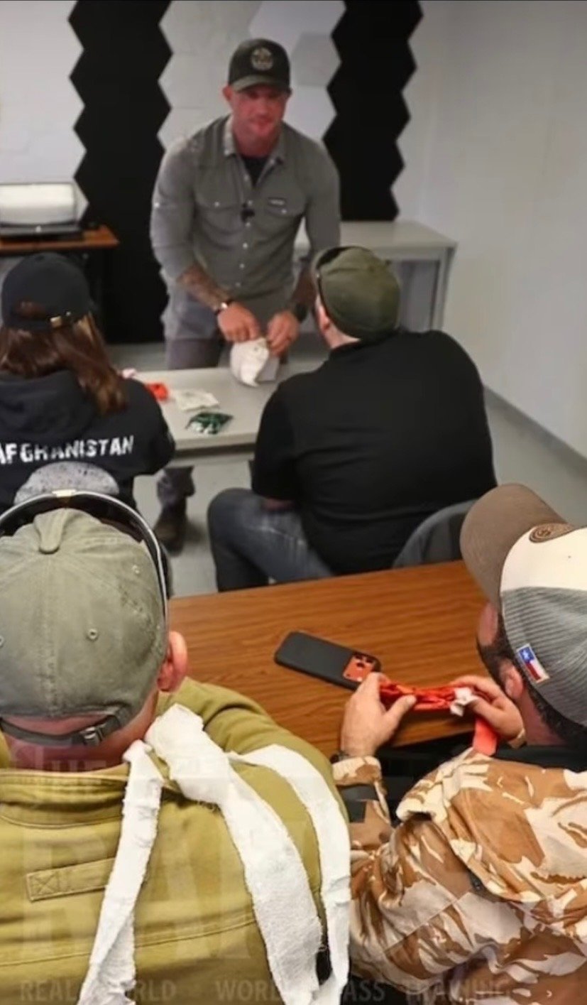 Man standing at a table in front of a seated group, holding a rolled cloth; the group of people is listening, some wearing caps and casual clothing, in a room with black soundproof panels on the wall.