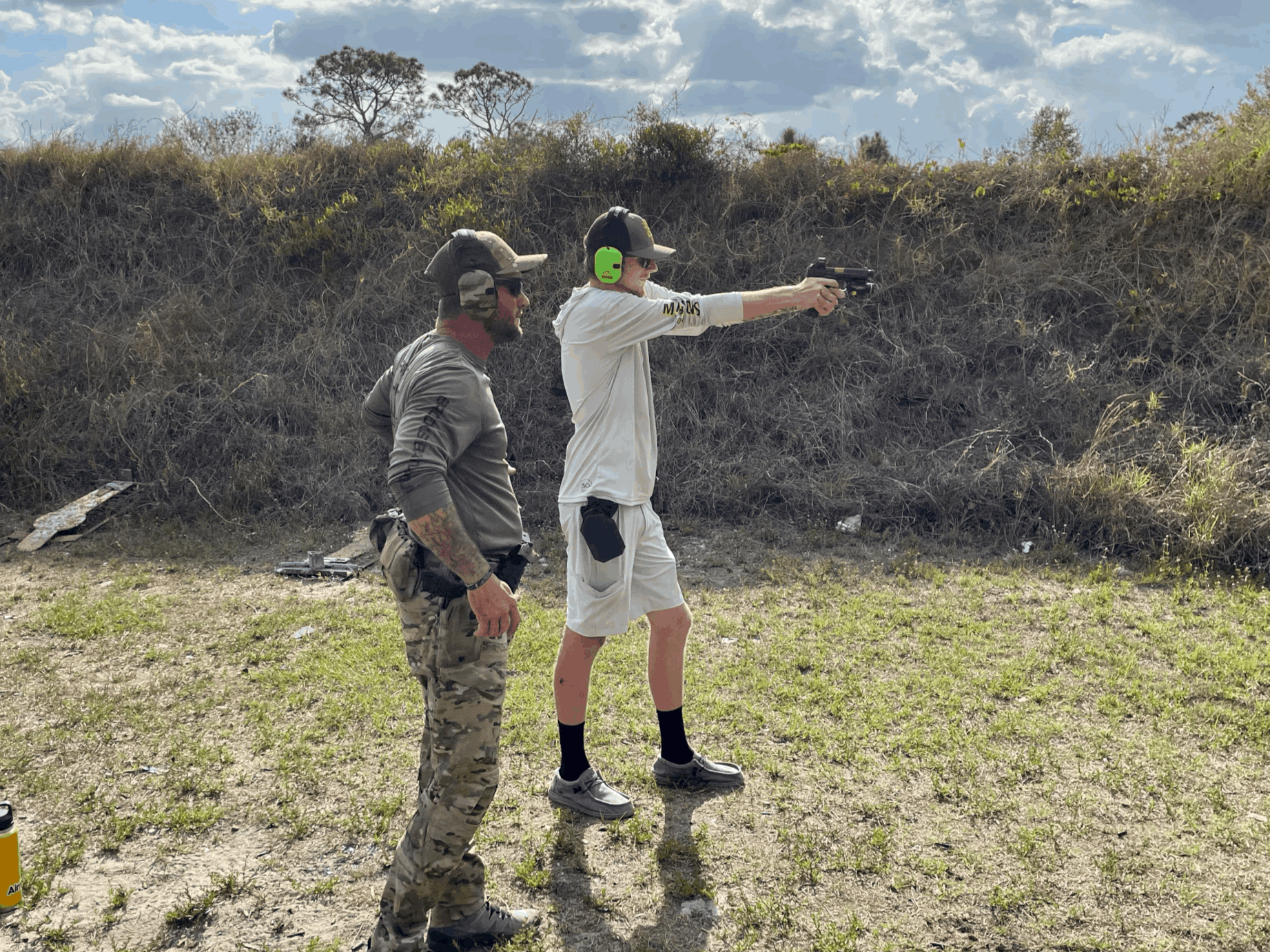 A man in camouflage pants and a gray shirt observing a young man in white shorts and a hoodie, who is aiming a pistol at a shooting range outdoors, with a dirt ground and a grassy hill in the background.