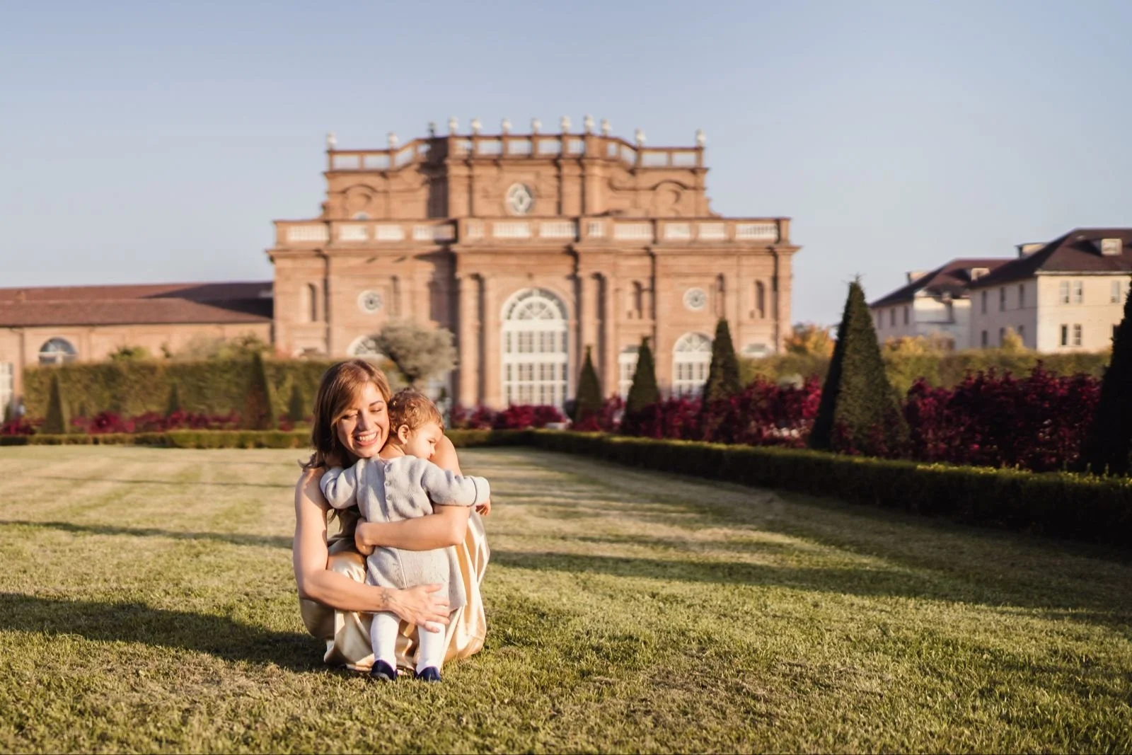 A woman and a young girl hugging in a garden with a large historic building in the background.