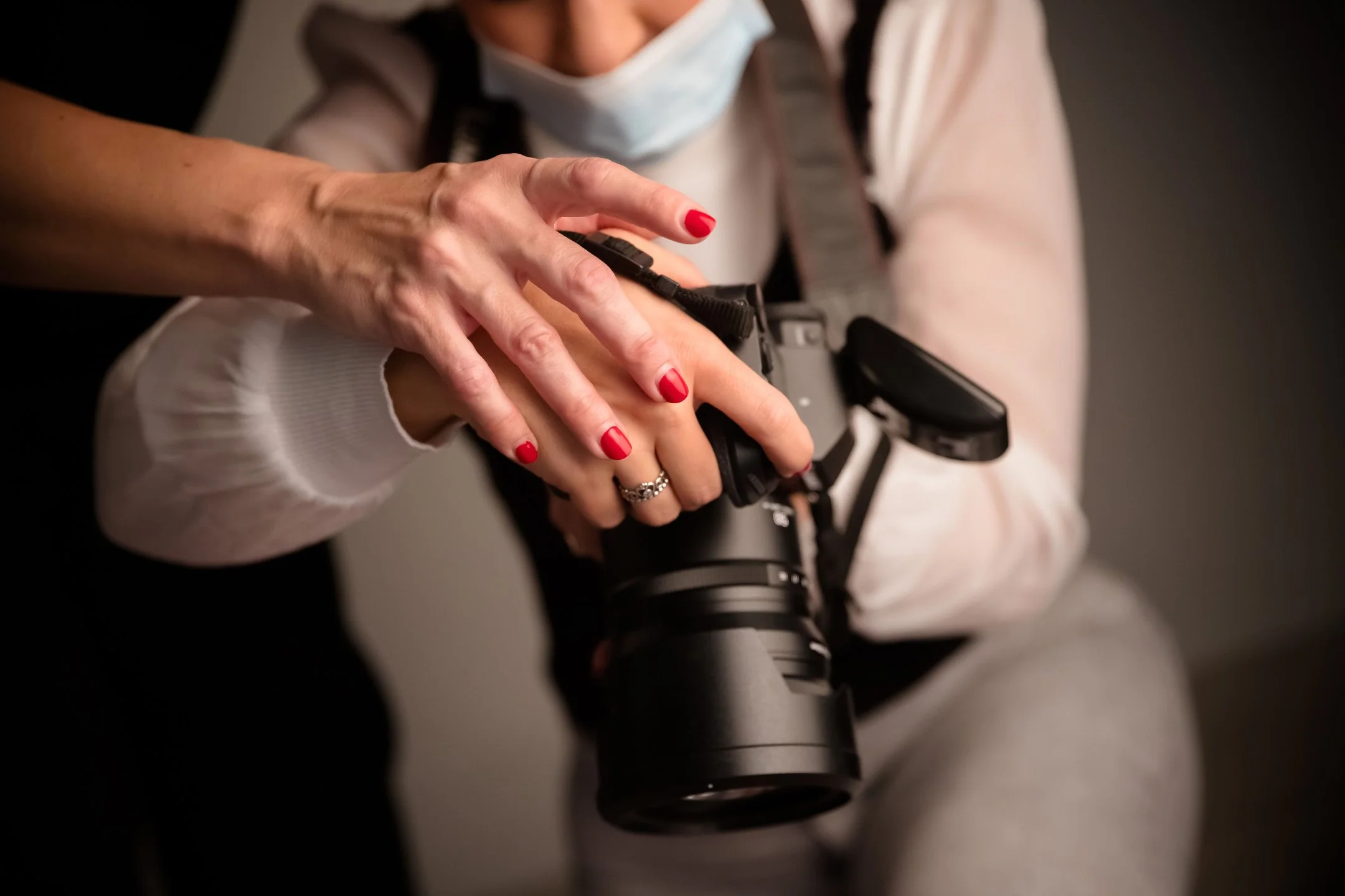 Person holding a camera with their right hand while their left hand is on the camera body, red nail polish, wearing a white long sleeve shirt, a face mask hanging on the neck, and a ring on the left hand.
