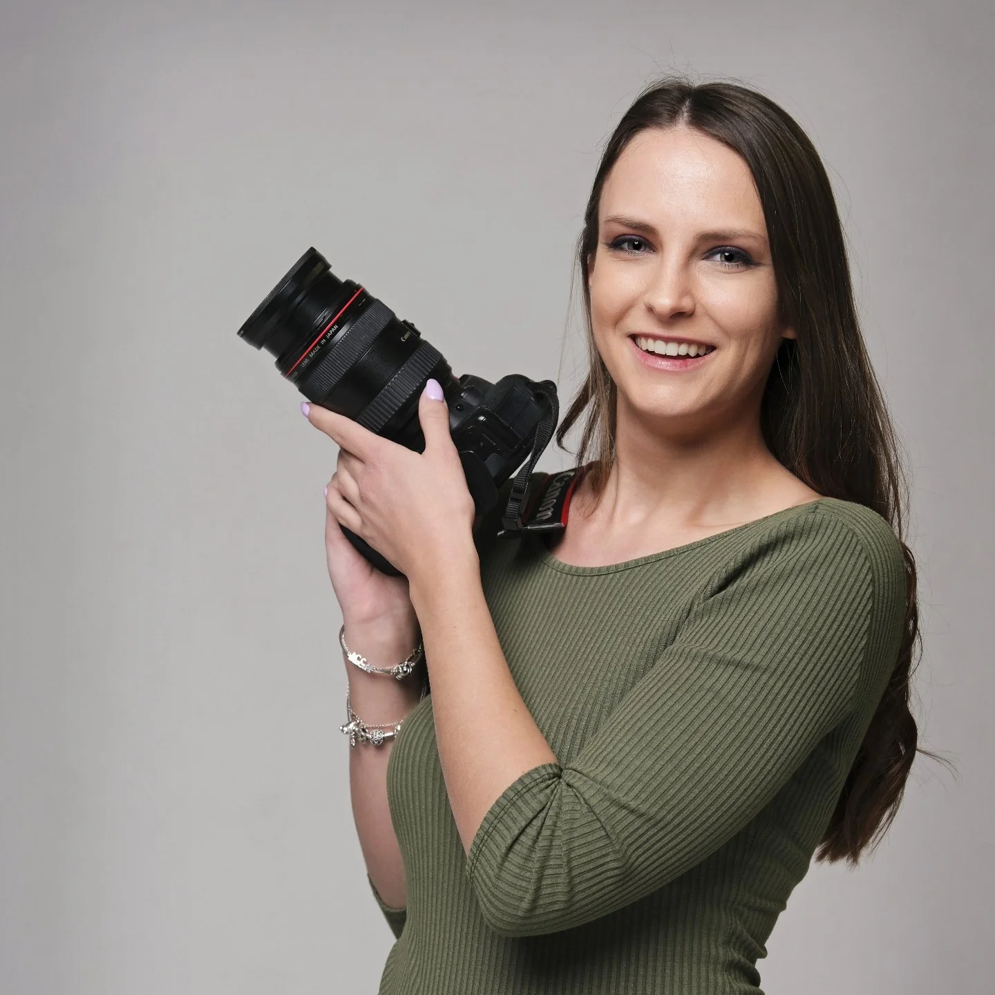 A young woman with long brown hair, wearing a green ribbed long sleeve shirt, holding a professional camera with a large lens, smiling at the camera against a plain gray background.