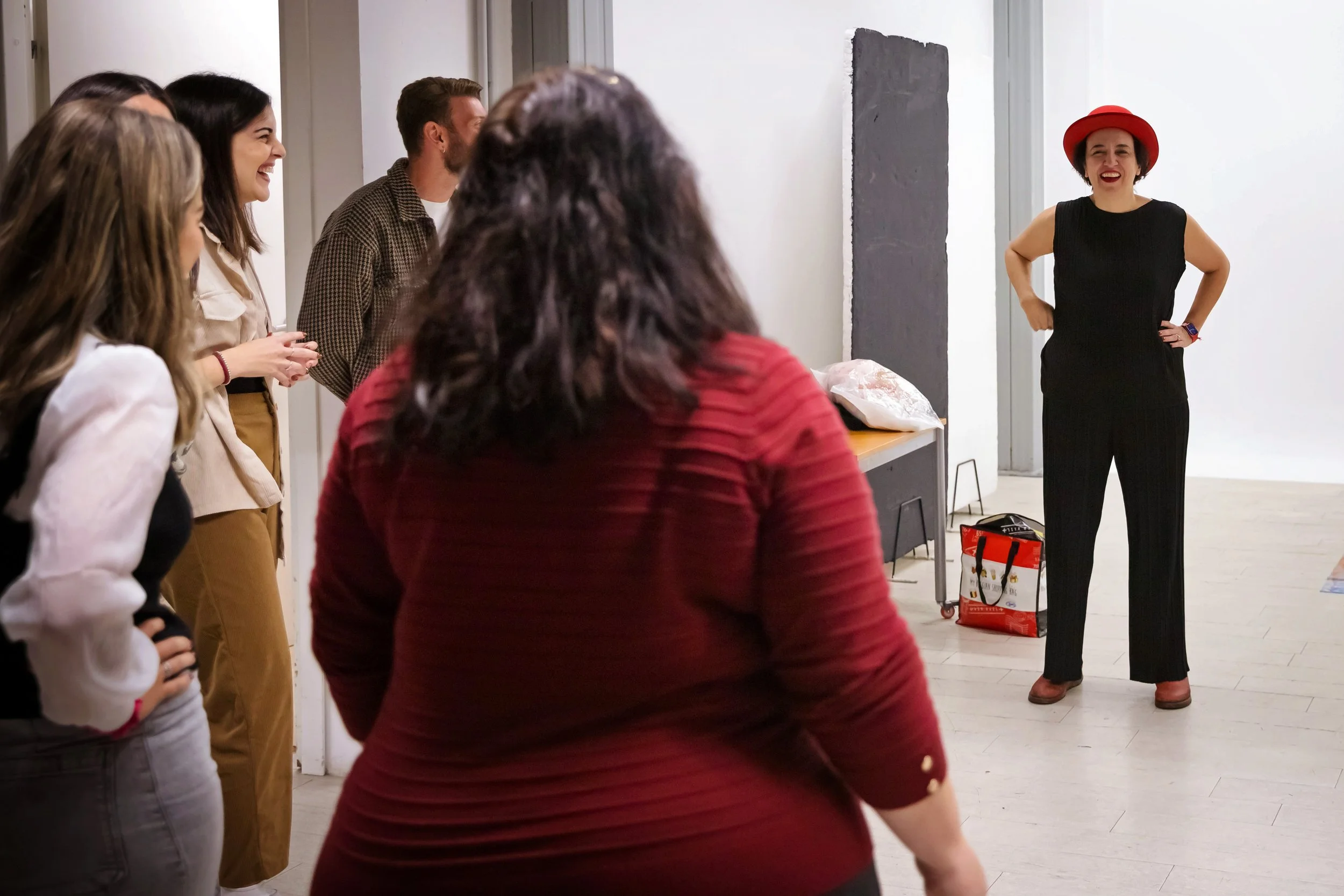 A woman in black pants and a red hat is performing or speaking to a group of diverse people in a room with white walls, a table, and miscellaneous items.