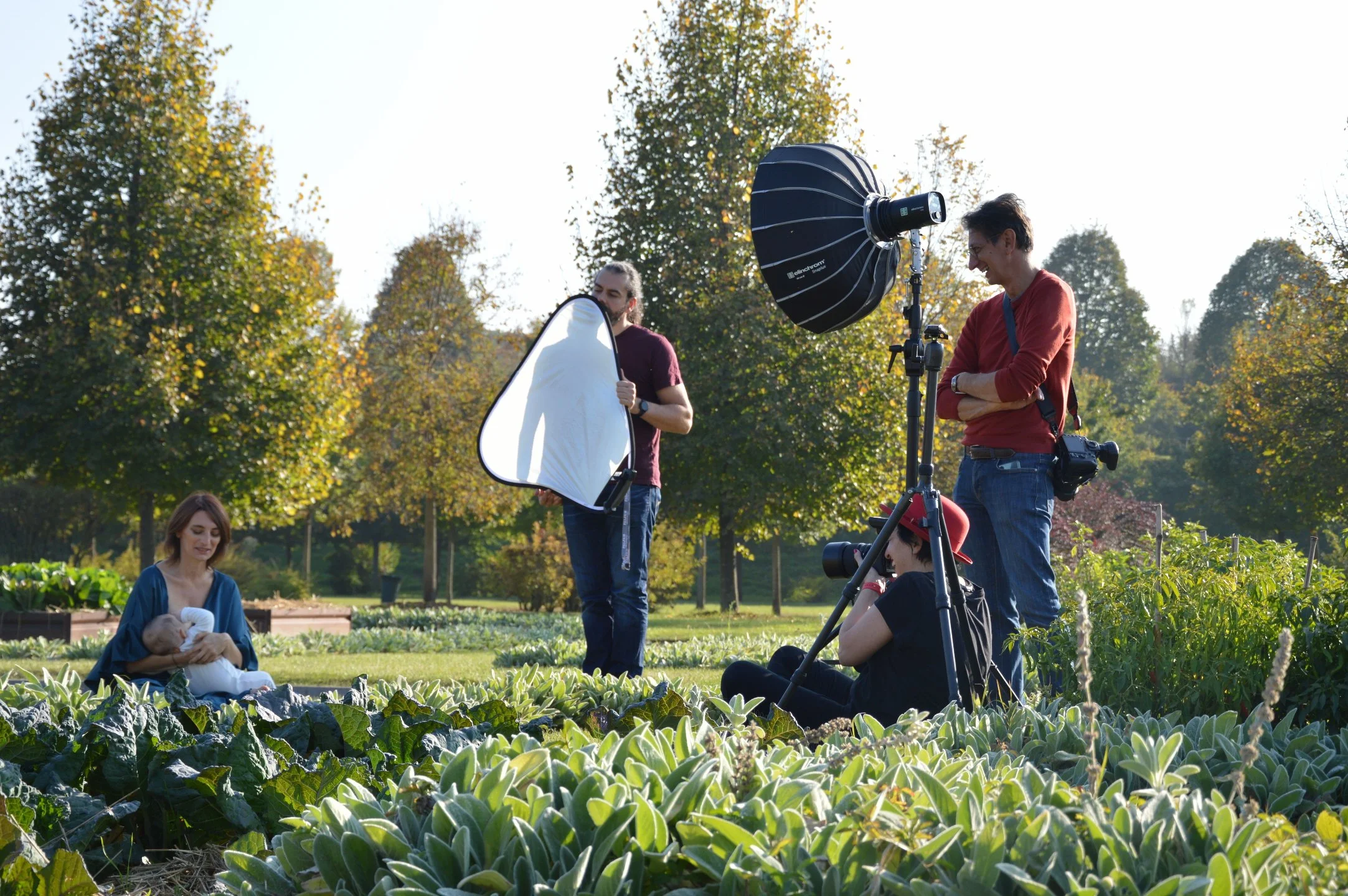 A photoshoot being conducted in a garden on a sunny day. A woman is holding a baby while sitting on the grass, surrounded by green plants. Two men and a woman are setting up photography equipment, including a large softbox and a camera on a tripod.