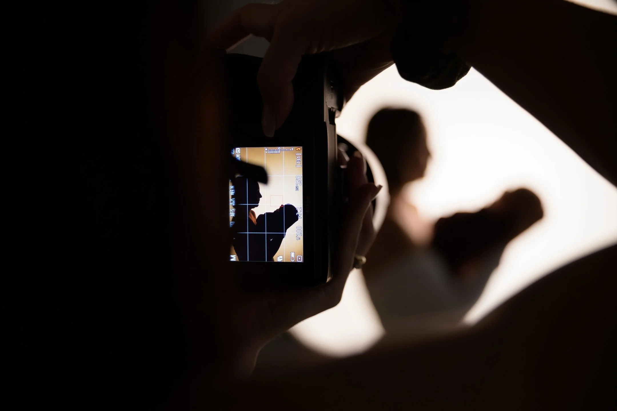 A photographer takes a picture of a woman holding a baby, with the image framed on a camera viewfinder showing their silhouette against a bright background.