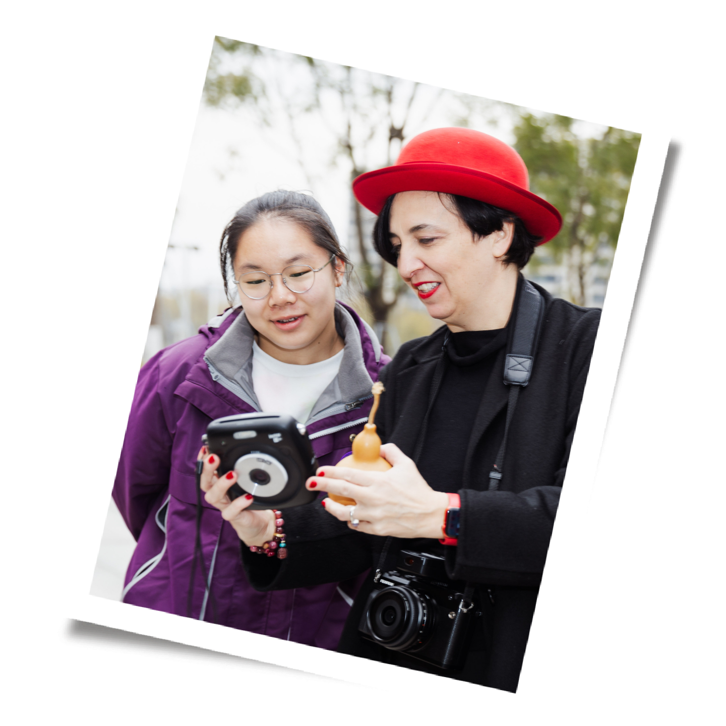 Two women looking at a device outdoors, one wearing a purple jacket and glasses, the other wearing a black jacket and red hat, holding a camera and a small radio.