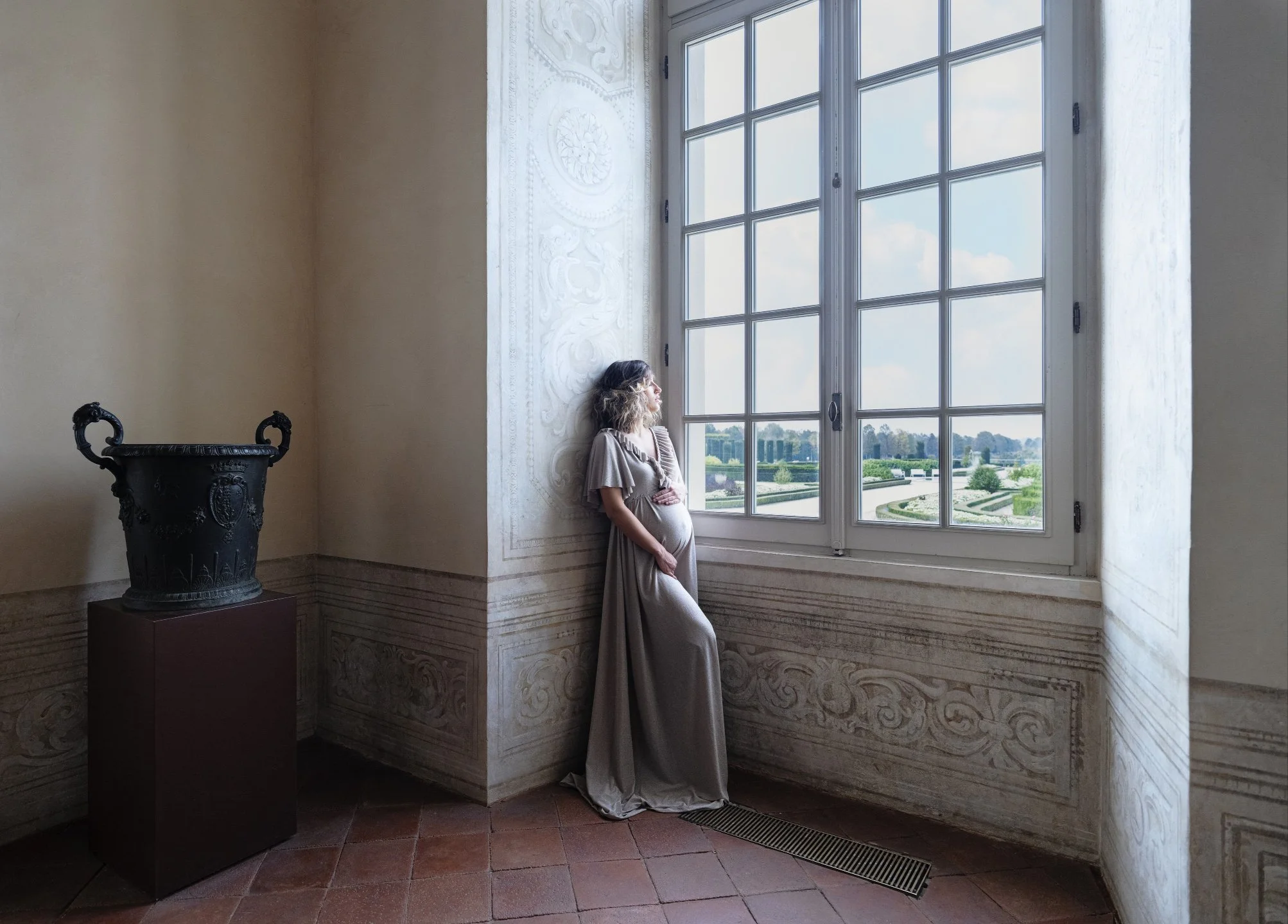 A pregnant woman with curly hair wearing a beige dress, standing in a room with large windows, looking outside at a scenic view with blue sky and green landscape.